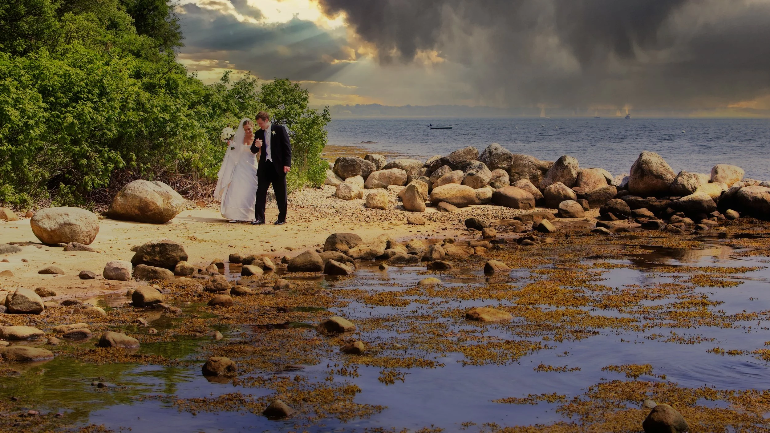 Bride and groom walking along a rocky Connecticut coastline as sailboats sail through stormy skies during a New London County wedding portrait.