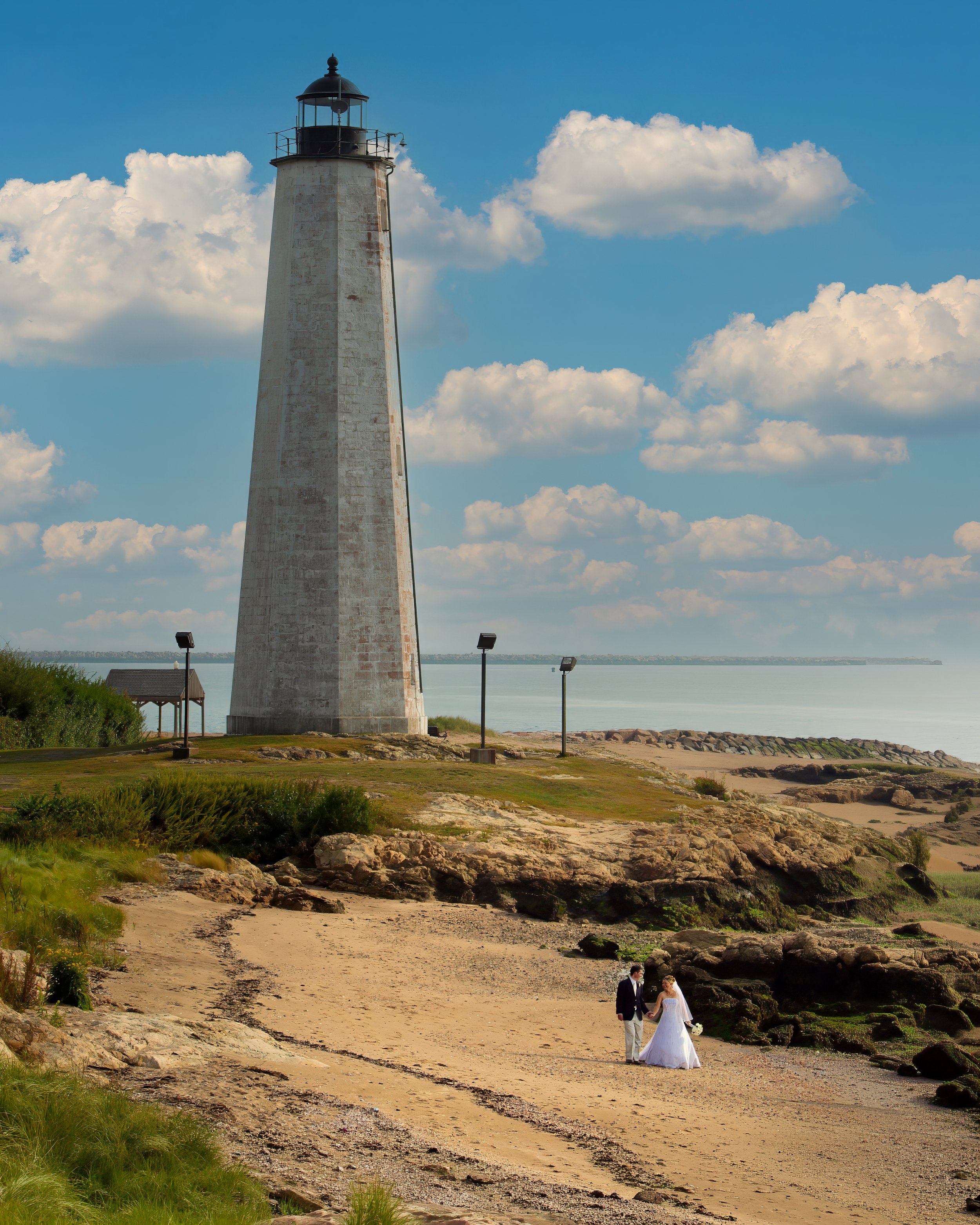 Bride and groom walking on the beach beneath the lighthouse at Lighthouse Point Park in New Haven Connecticut