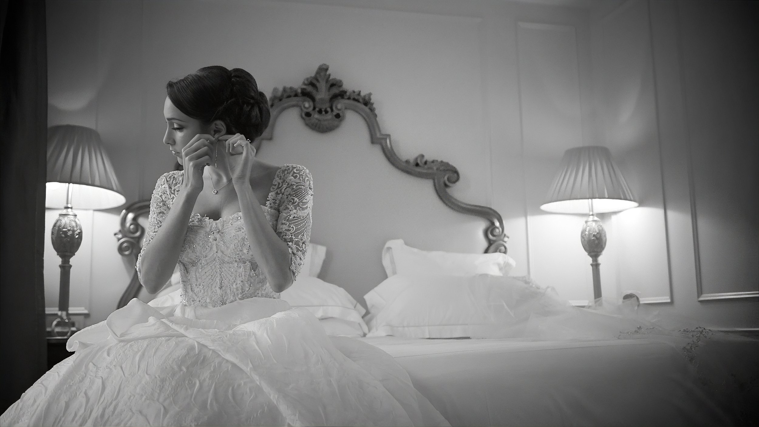 Bride sitting on the edge of her bed putting on earrings in her Plaza Hotel NYC bridal suite.