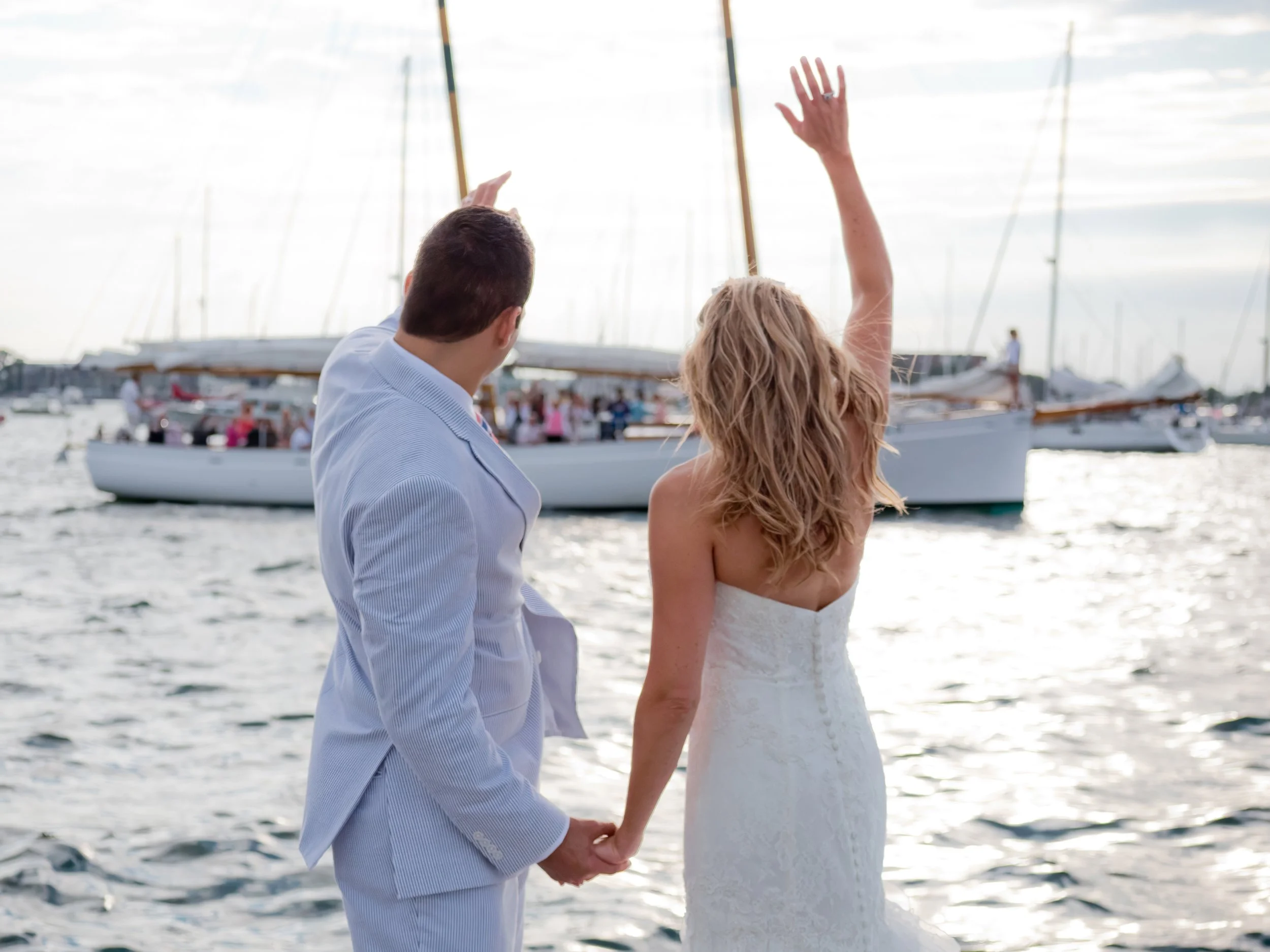 Bride and groom wave to a passing boat at the Newport Yachting Center harbor during their wedding day.