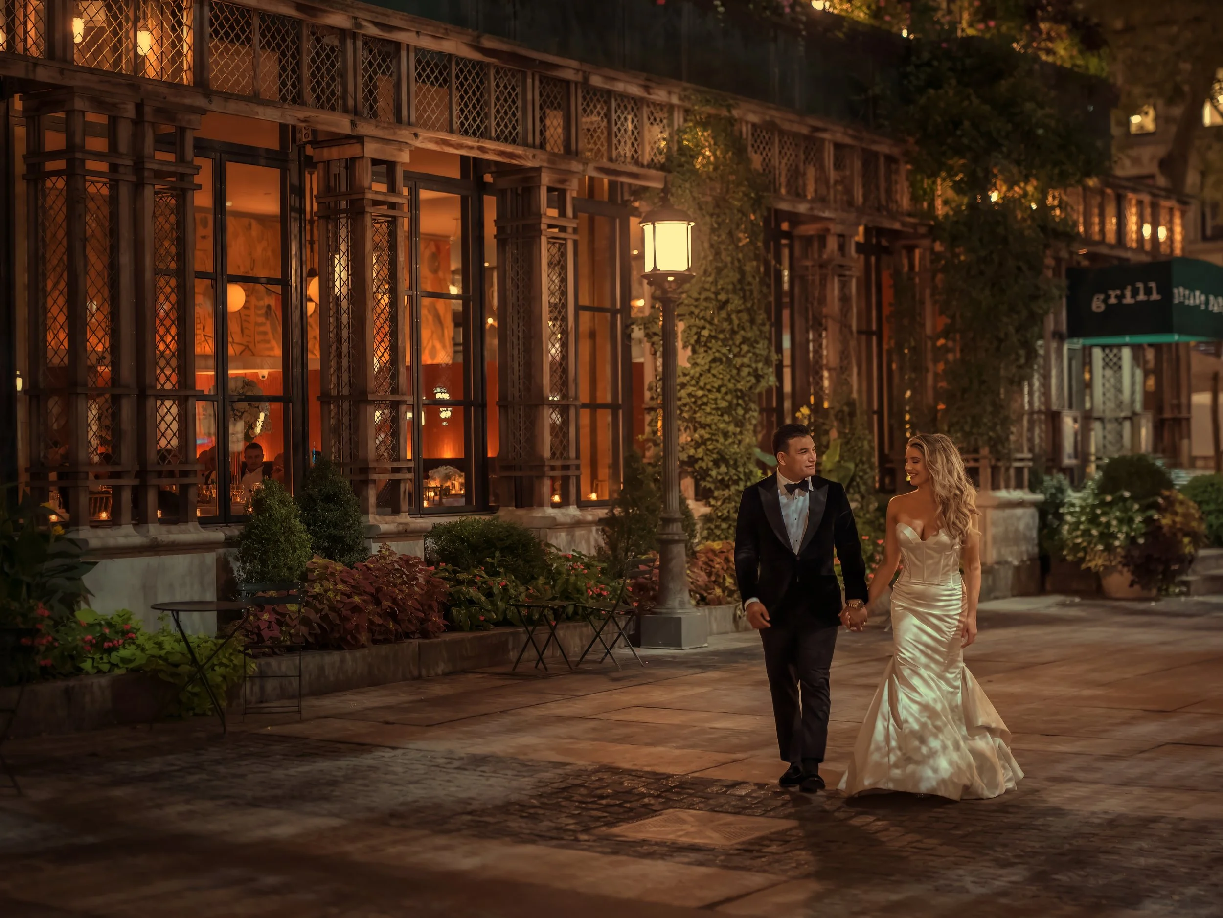 Bride and groom walk hand in hand through Bryant Park Grill at night, surrounded by romantic lighting and the glow of Midtown Manhattan.