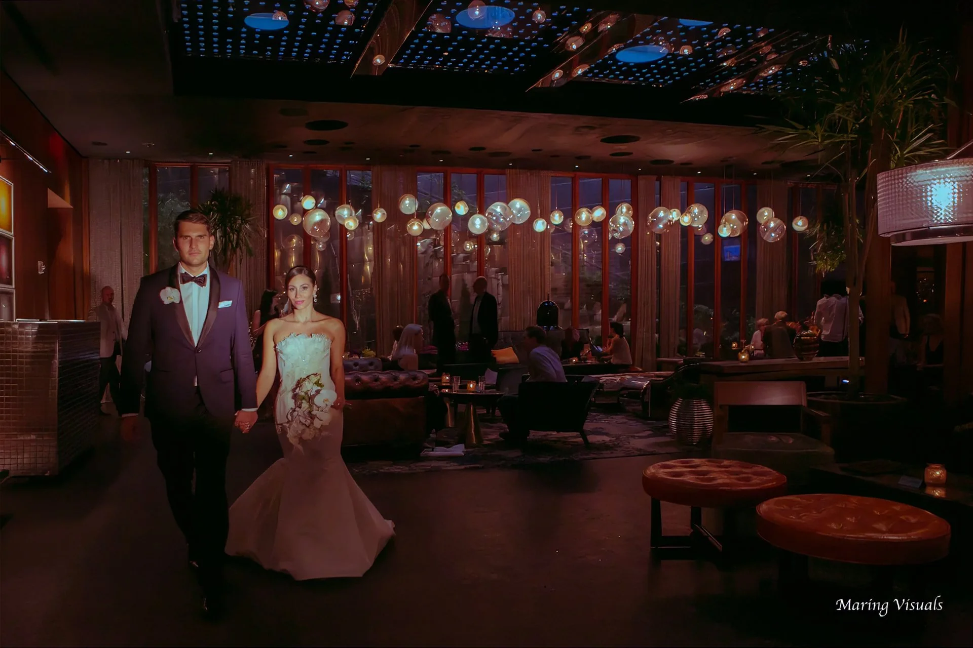 Bride and groom walk hand in hand through a dramatically lit contemporary lounge following their wedding celebration.