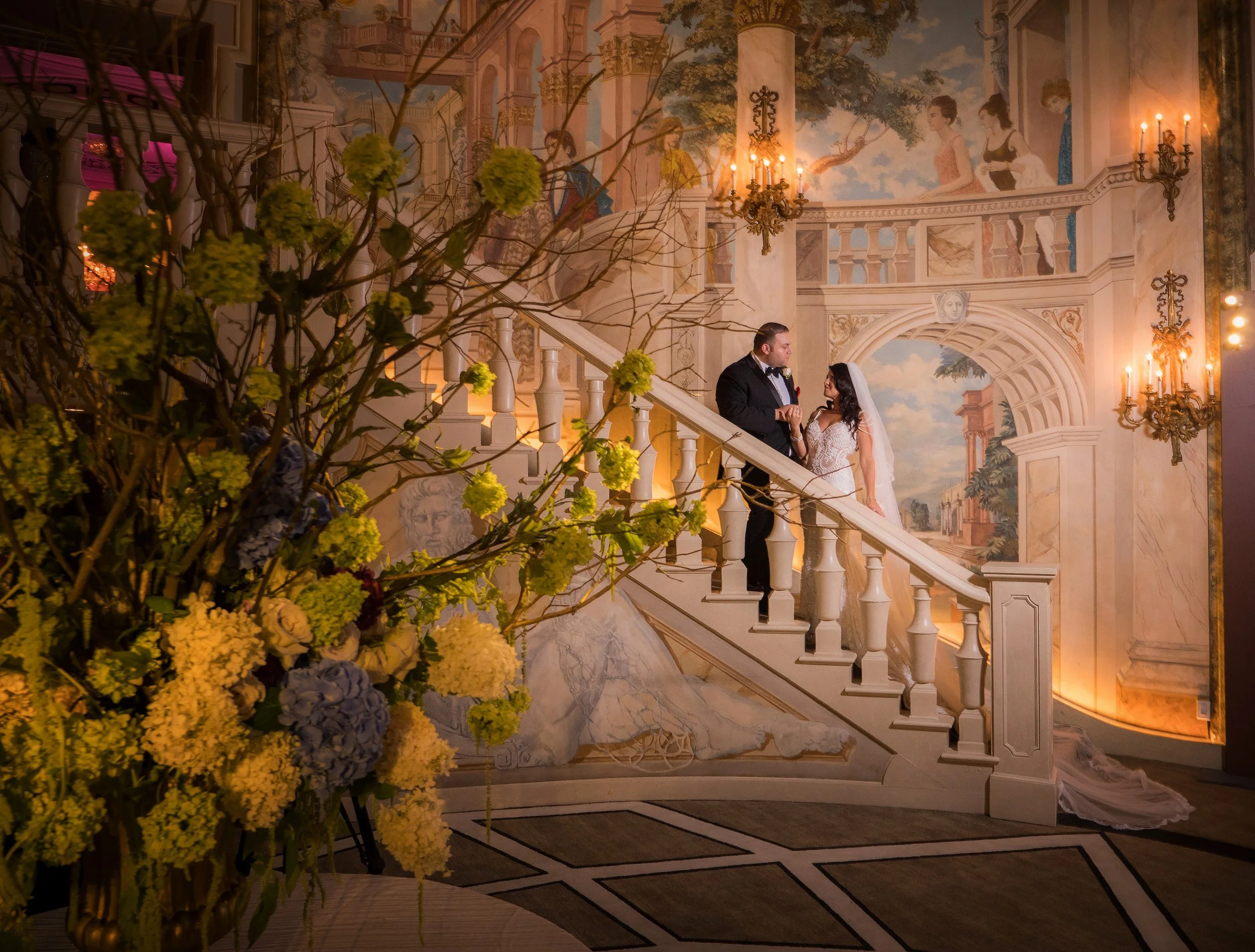 Bride and groom pose for a romantic portrait on the Rotunda Room staircase at The Pierre Hotel NYC, framed by floral arrangements