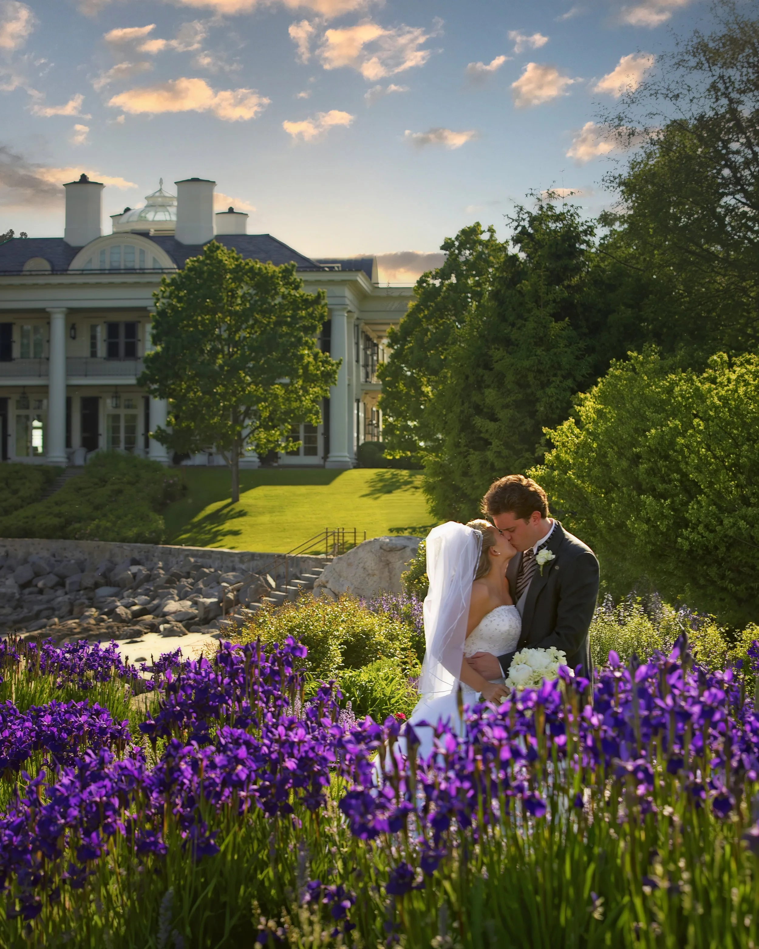 Bride and groom kiss on the grounds of Belle Haven Yacht Club surrounded by blooming flowers.