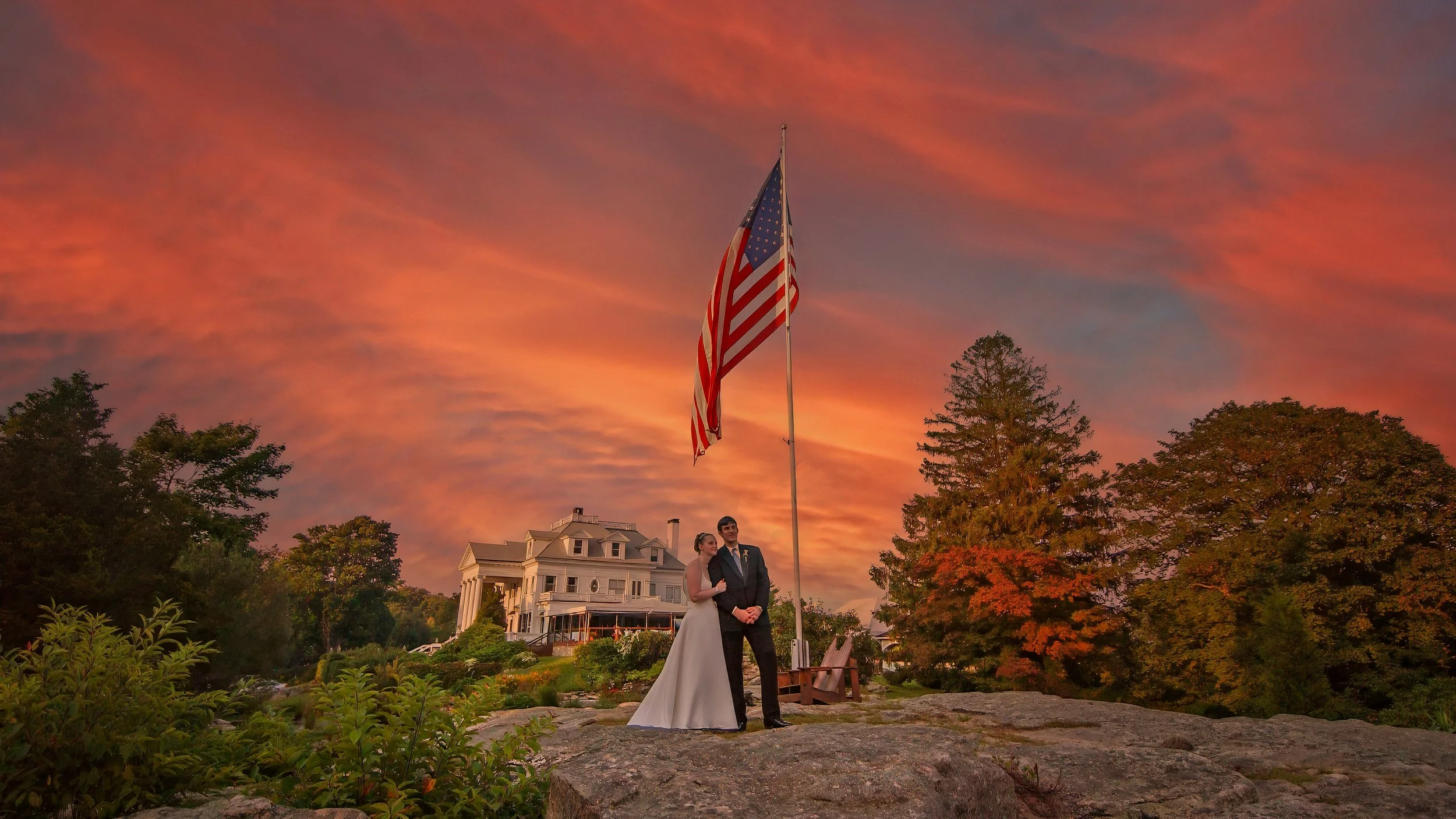 Bride and groom beneath American flag at sunset with Haley Mansion backdrop during Inn at Mystic wedding Connecticut
