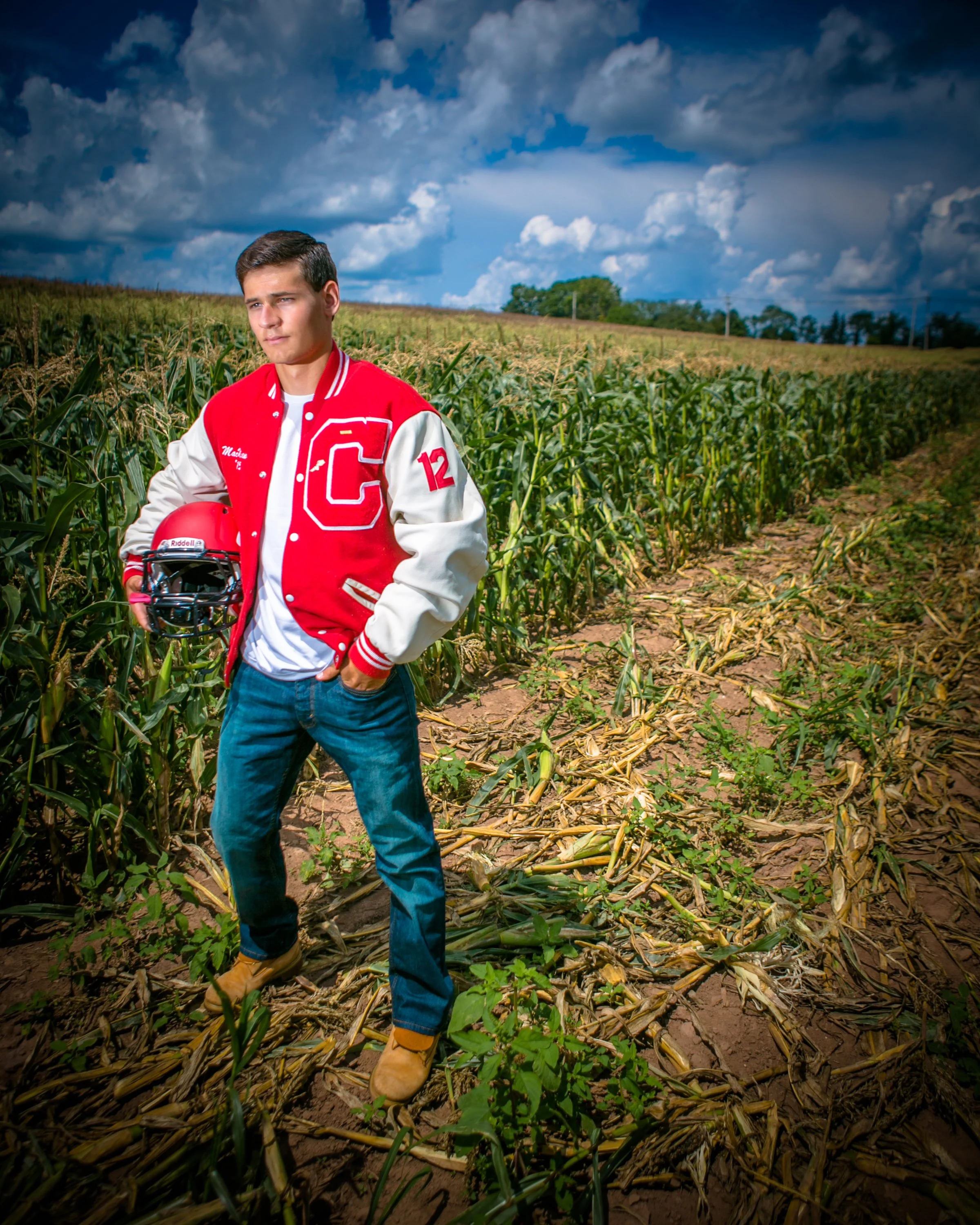 Senior portrait of a young man in a school jacket holding a football helmet in a cornfield.