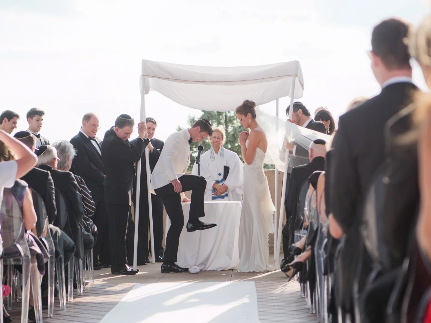 Groom breaks the glass beneath a chuppah as the bride and guests watch during an outdoor Jewish wedding ceremony.