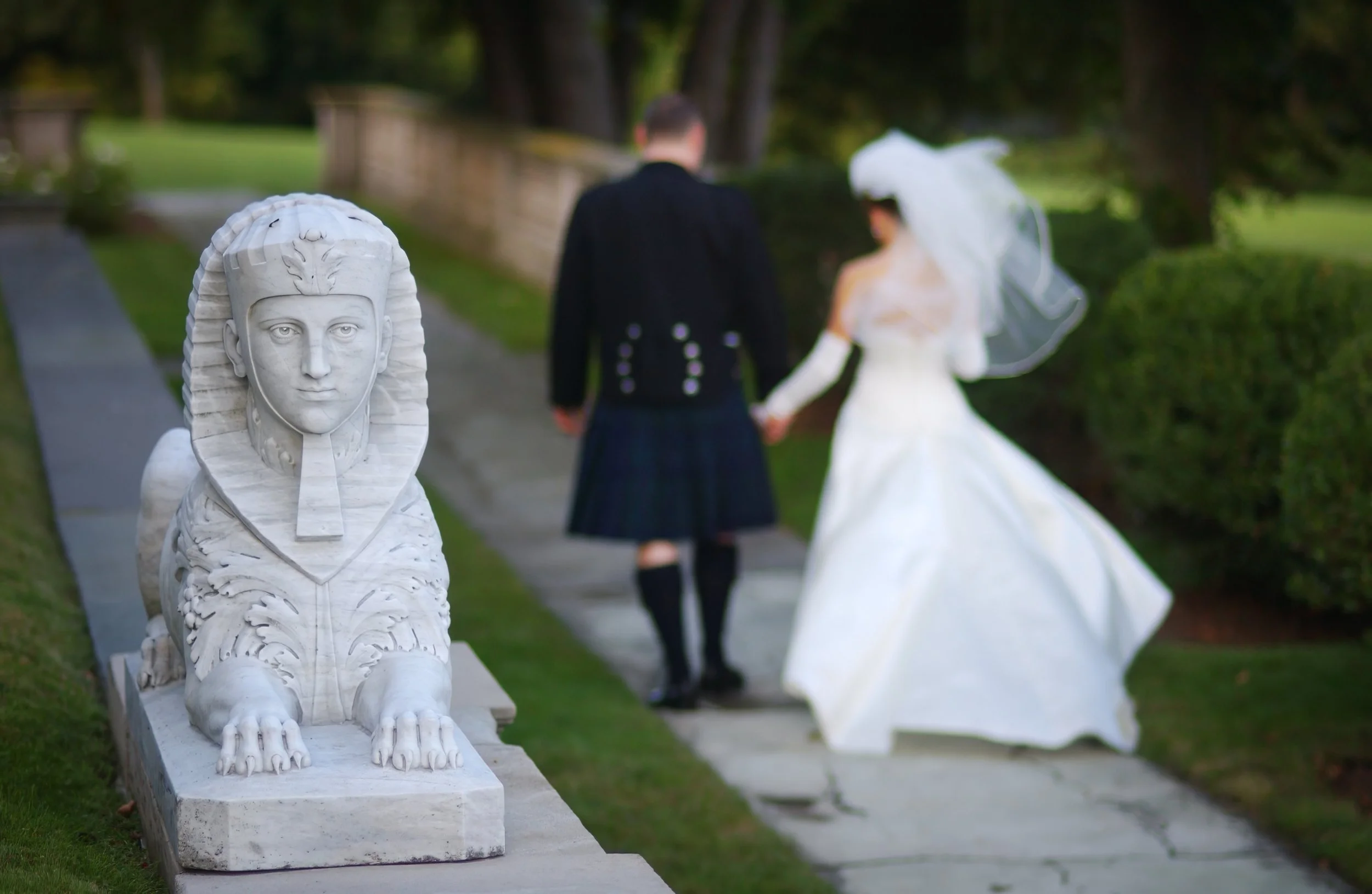 Bride and groom walk hand in hand down a stone path at Aldrich Mansion with statues along the path.