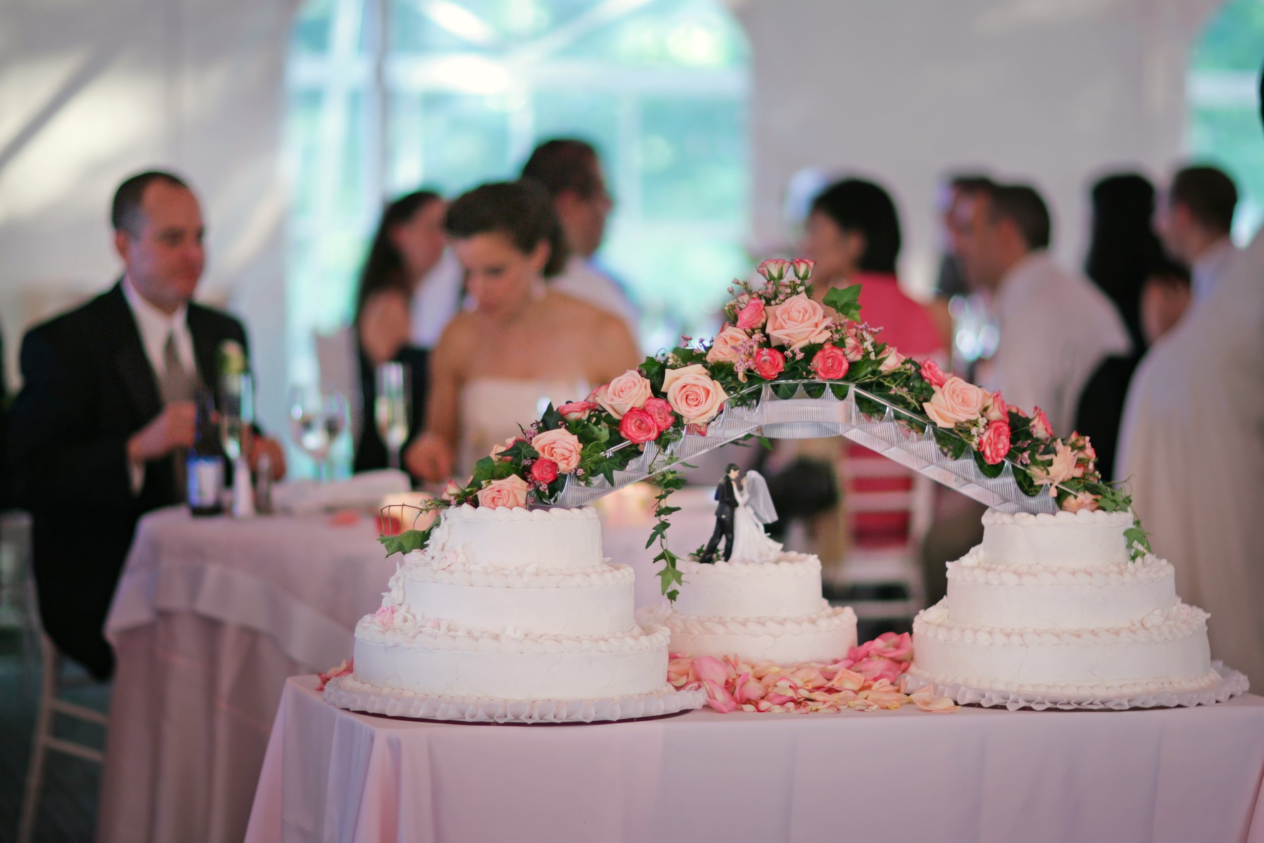 Wedding cake displayed under a tented reception as bride and groom enjoy dinner in the background.