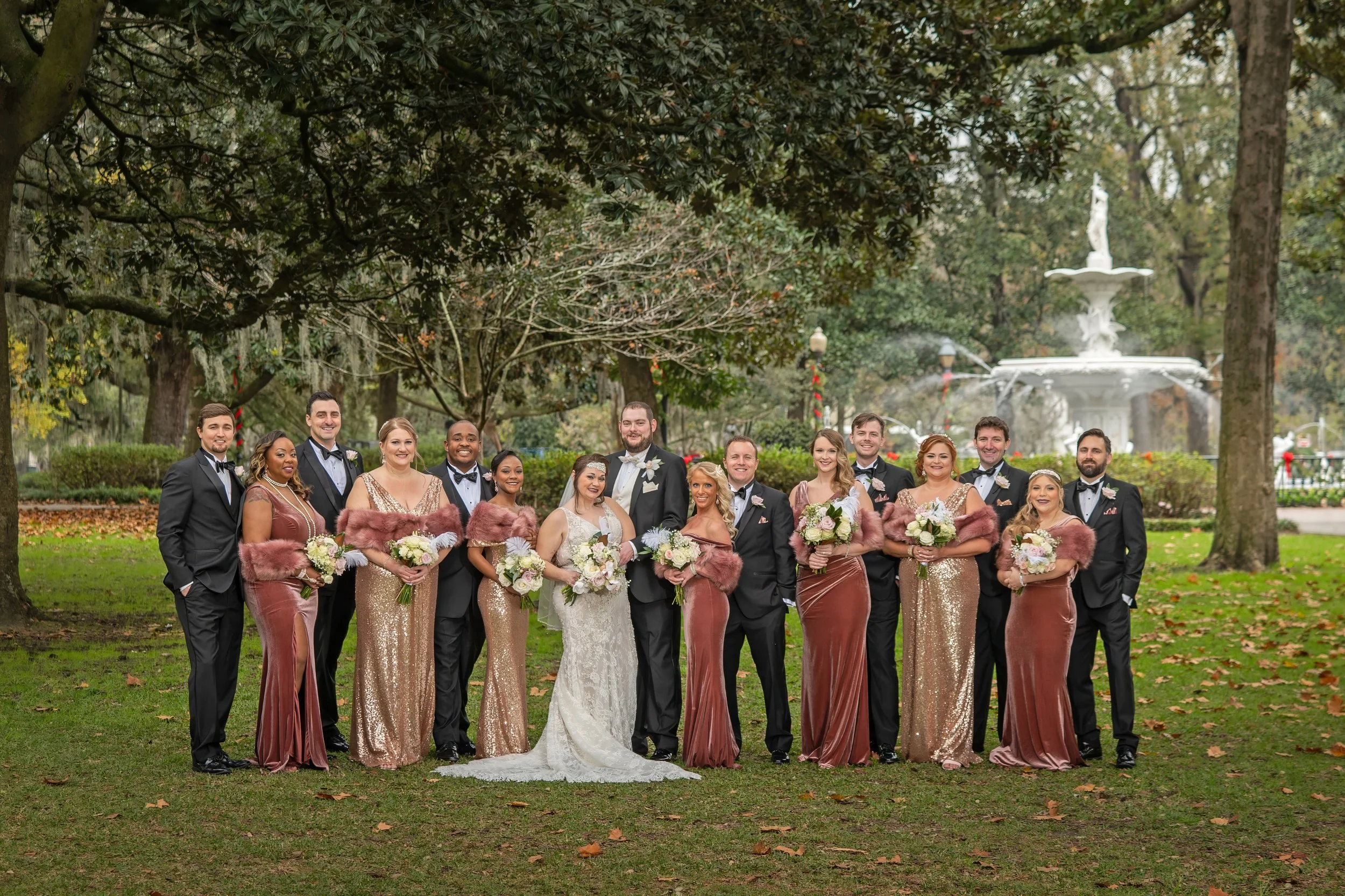 Large bridal party poses under Magnolia tree shade with Forsyth Park Fountain in the distance, capturing a joyful wedding portrait in Savannah.