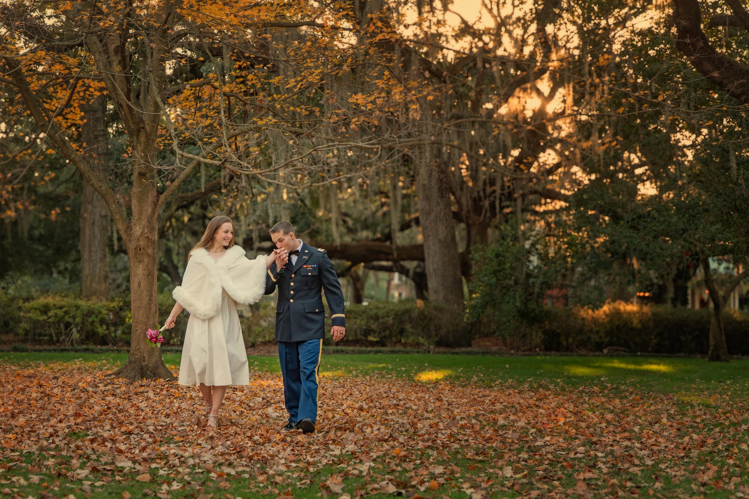 Groom kisses bride’s hand as they walk through fall leaves in Forsyth Park, Savannah, creating a romantic seasonal moment.