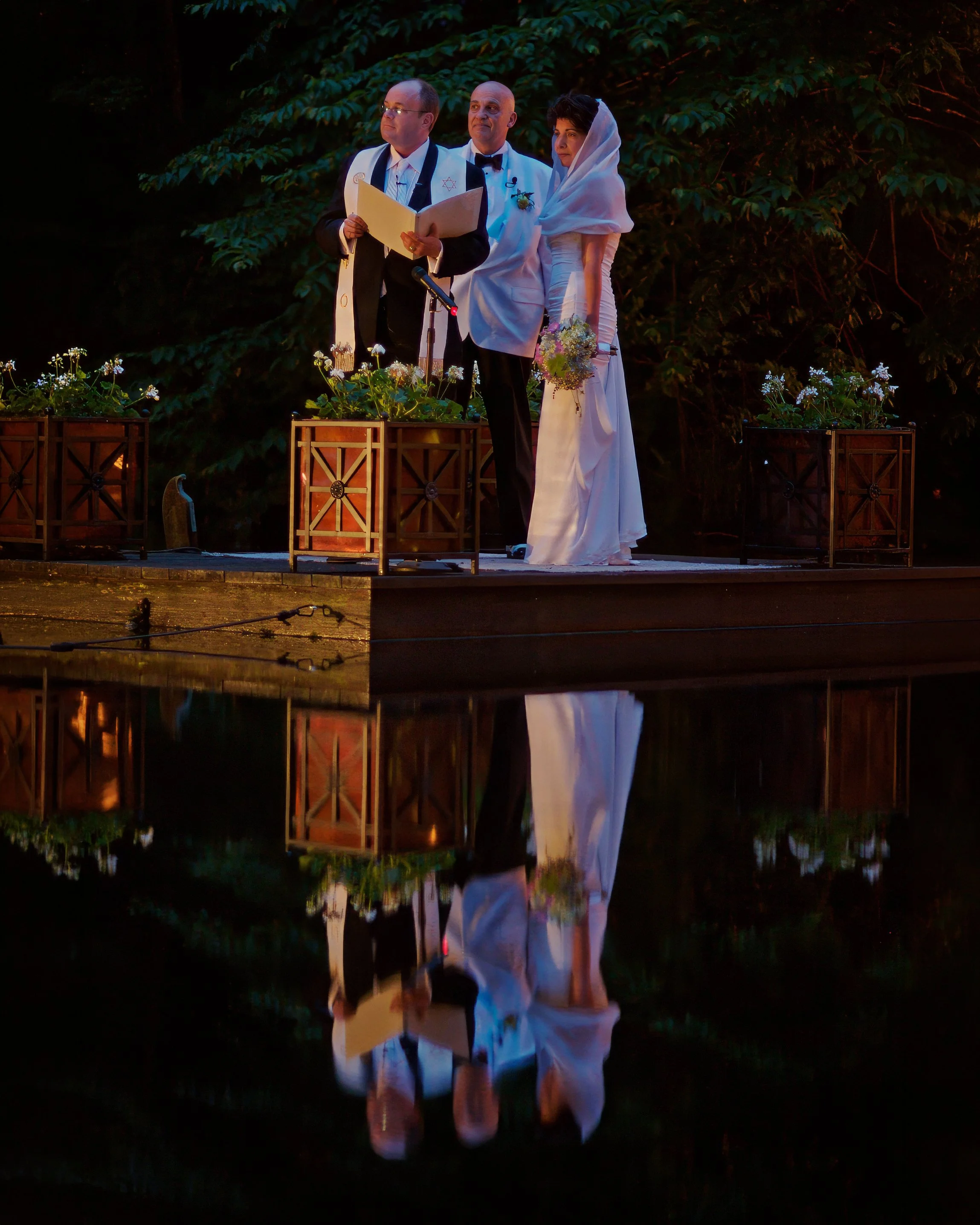 Bride and groom standing on a dock beside a reflective pond while violinists perform across the water at a Connecticut estate wedding.