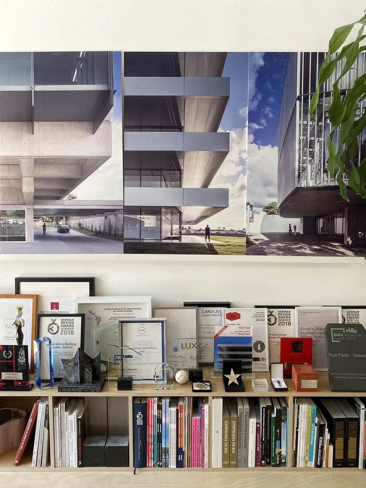 Display shelf with international architecture awards, certificates, and books beneath a triptych of modern architectural renderings at the studio of ruiz pardo – nebreda