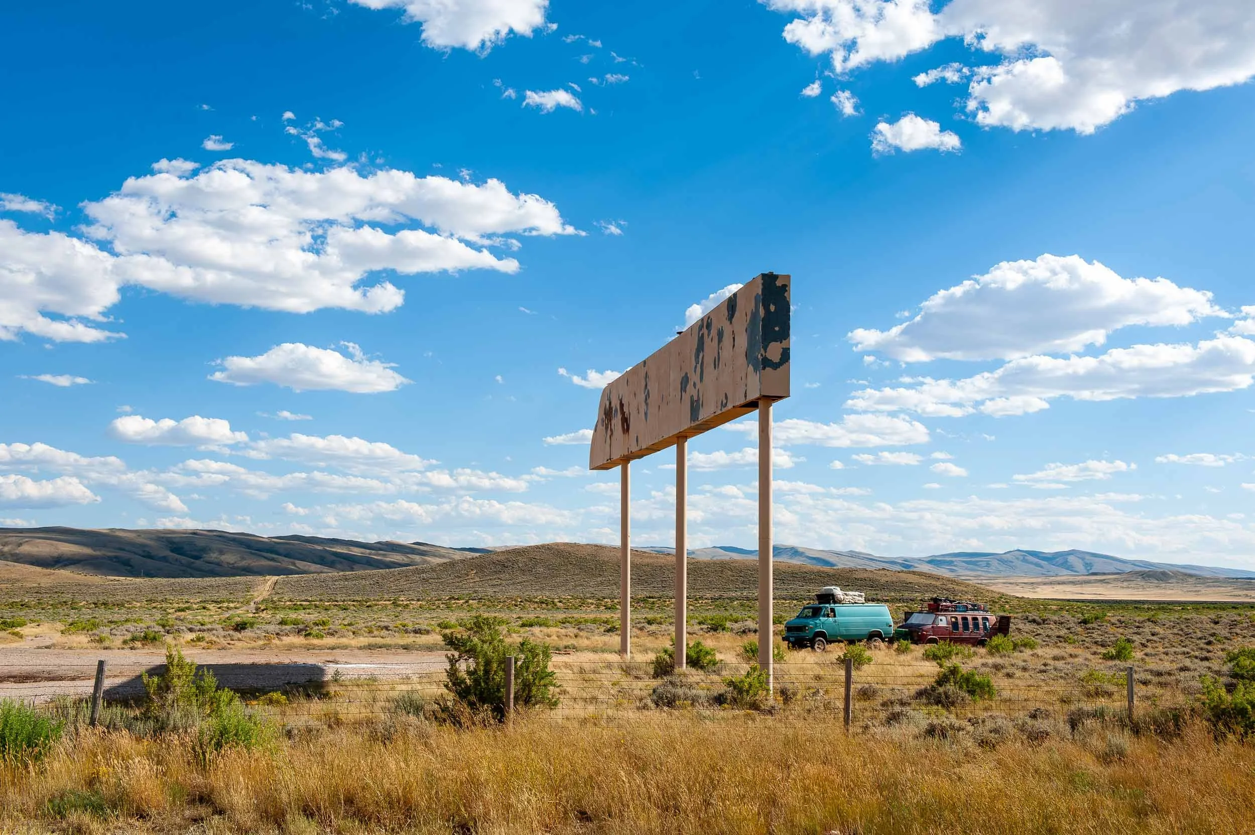 Empty billboard in a rural landscape with two parked camper vans, surrounded by dry grass and sparse bushes, under a bright blue sky with scattered white clouds.