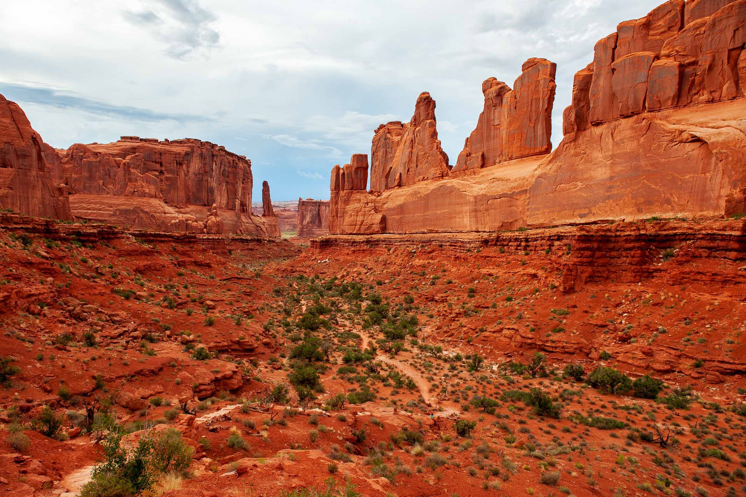 Red rock canyons and formations in a desert landscape with sparse green vegetation and cloudy sky.
