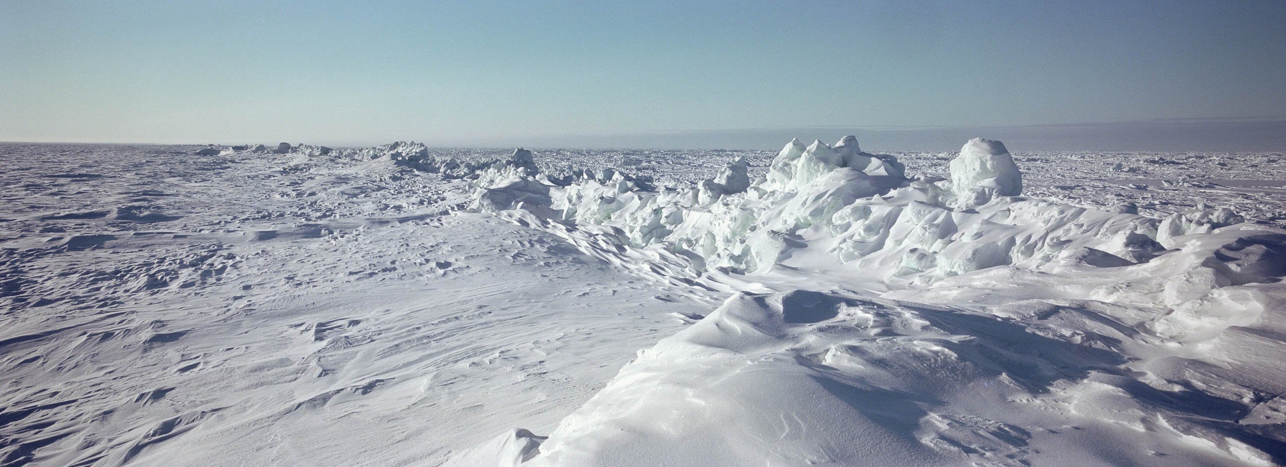 Expansive icy Arctic landscape with snow-covered ground and large ice formations under clear blue sky.