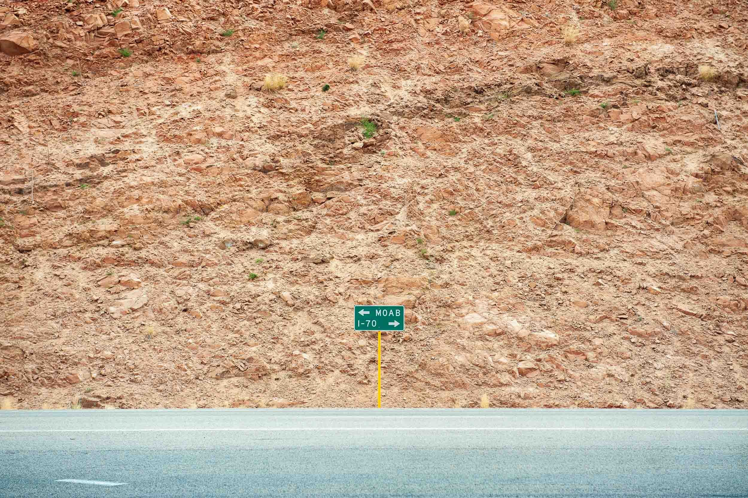 Road with a green directional sign indicating left to MOAB and I-70, and right to same destinations, against a rocky, reddish hillside.