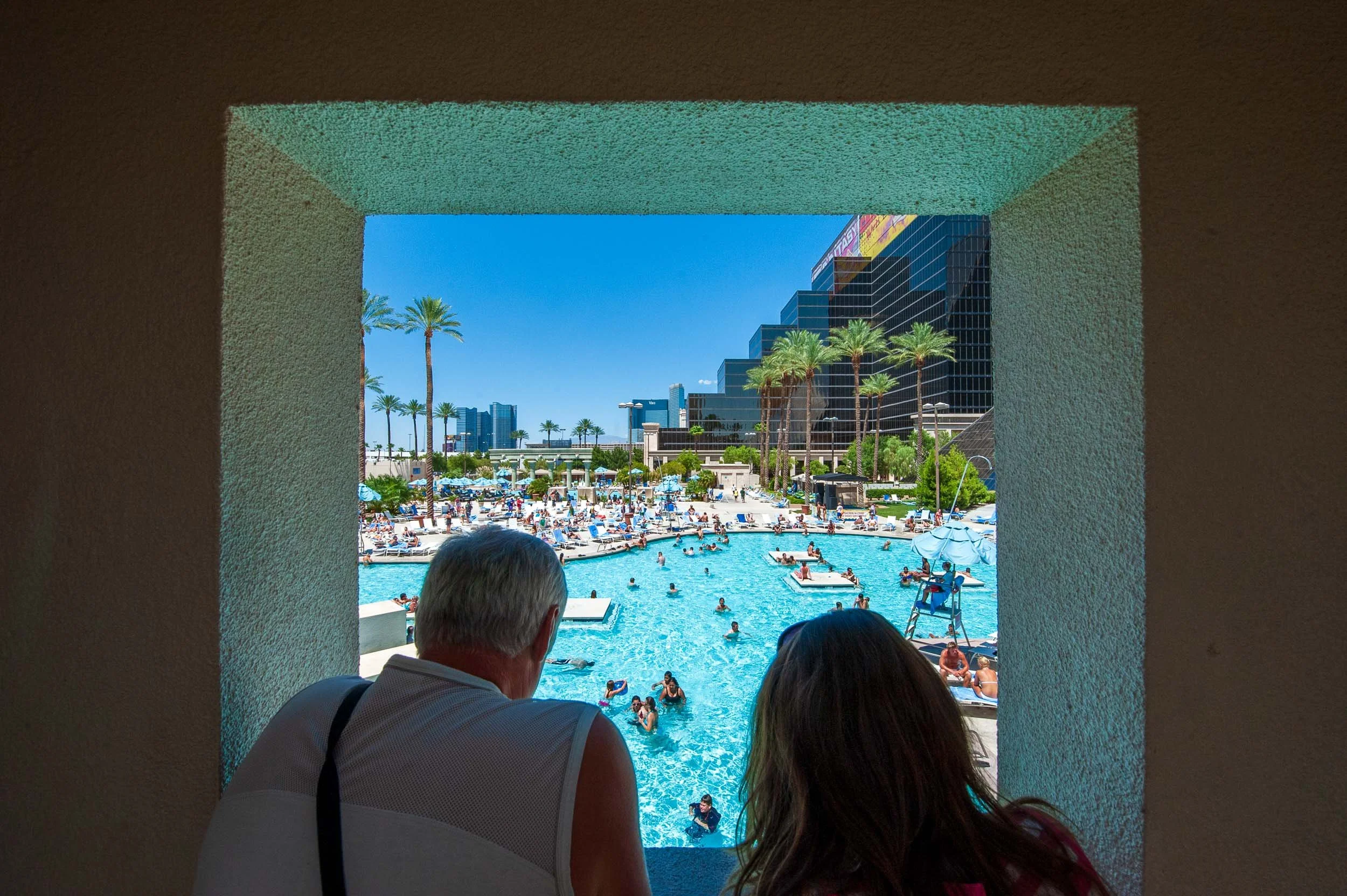 View of a busy hotel pool seen through a small window, with a man and woman sitting in the foreground.