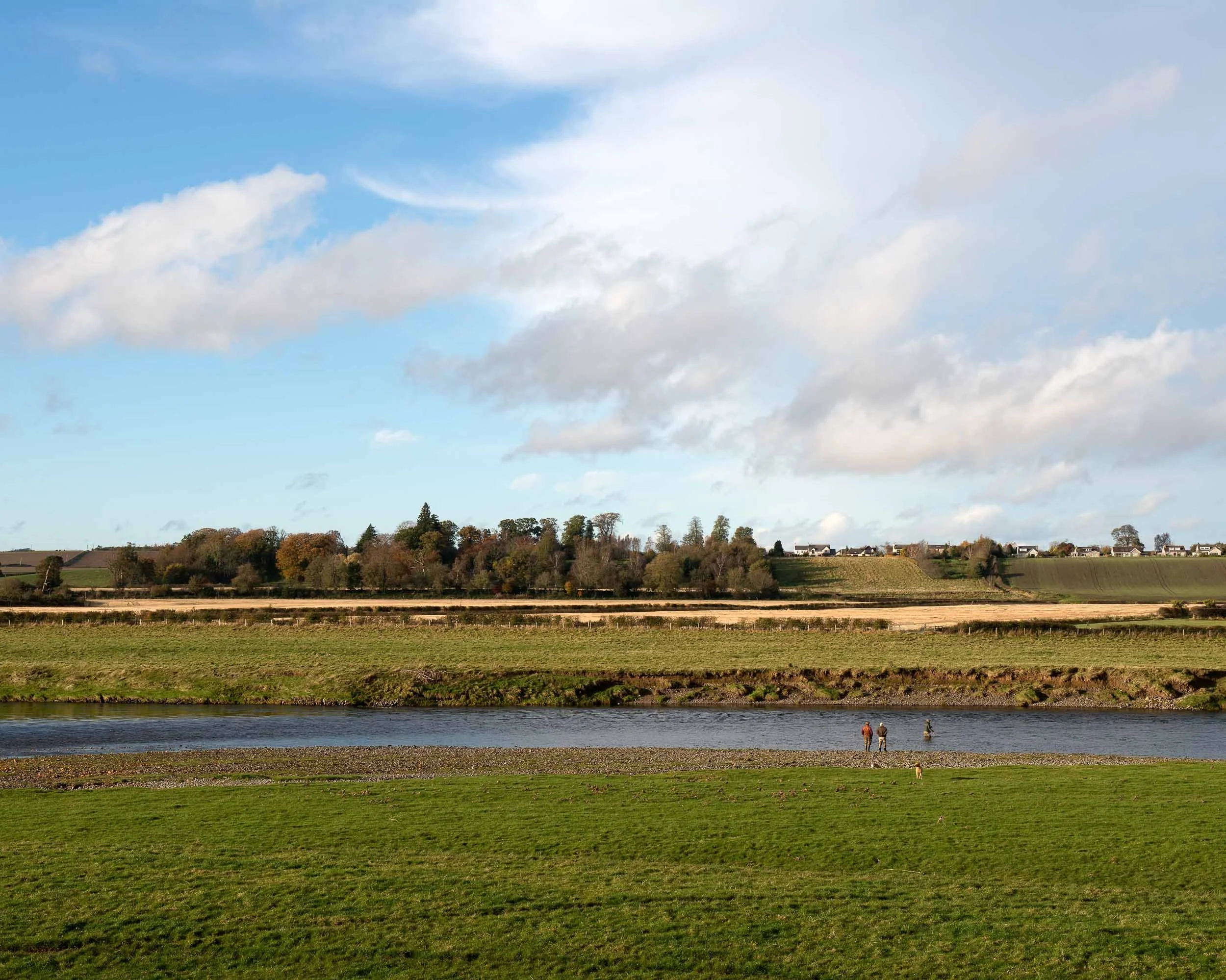 A scenic landscape with a river, green grassy fields, trees, and a clear sky with scattered clouds. There are three people near the riverbank, one with a dog.