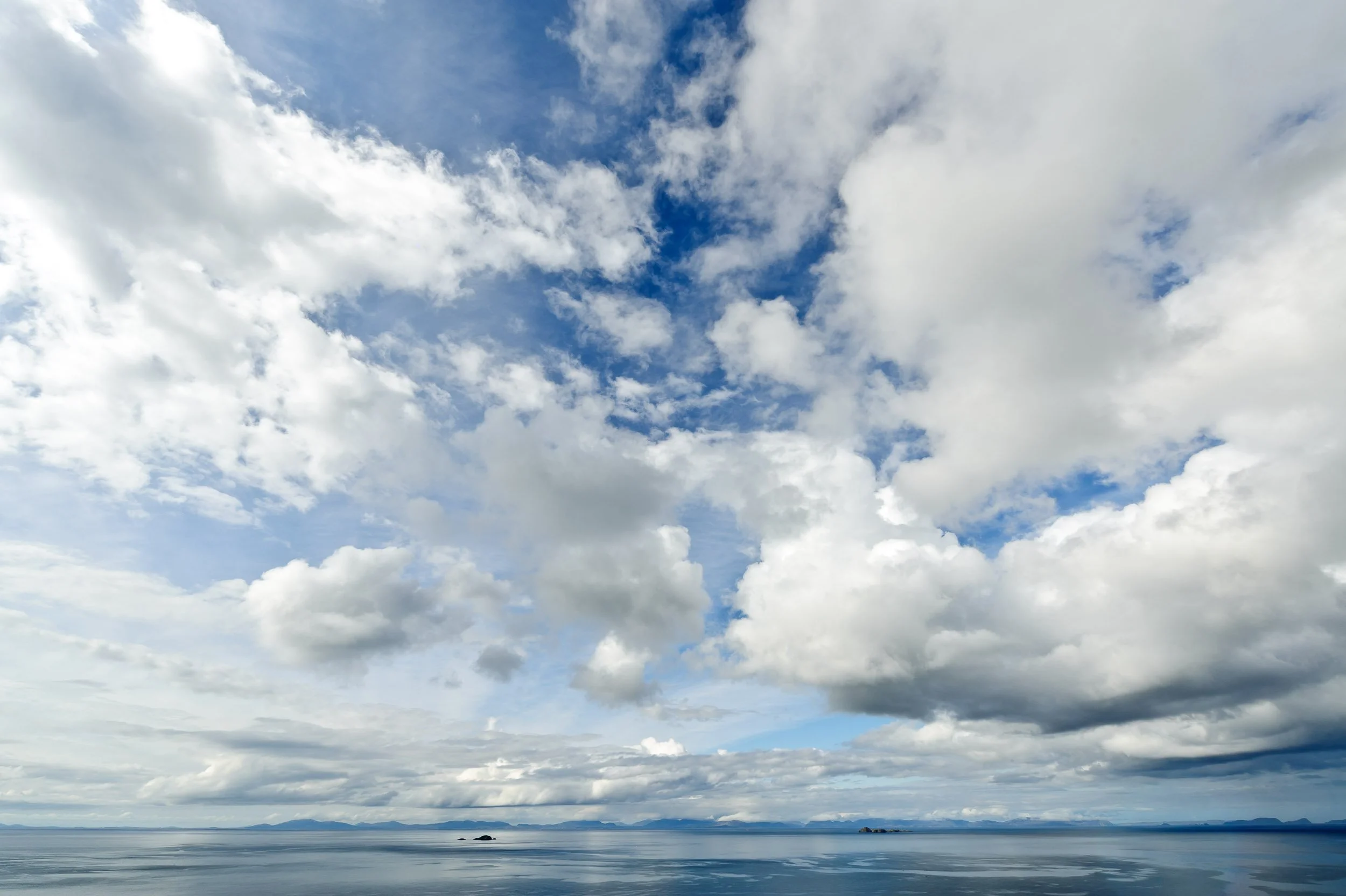 A vast body of water under a partly cloudy sky with various clouds, some darker, and a distant horizon of mountains or islands.