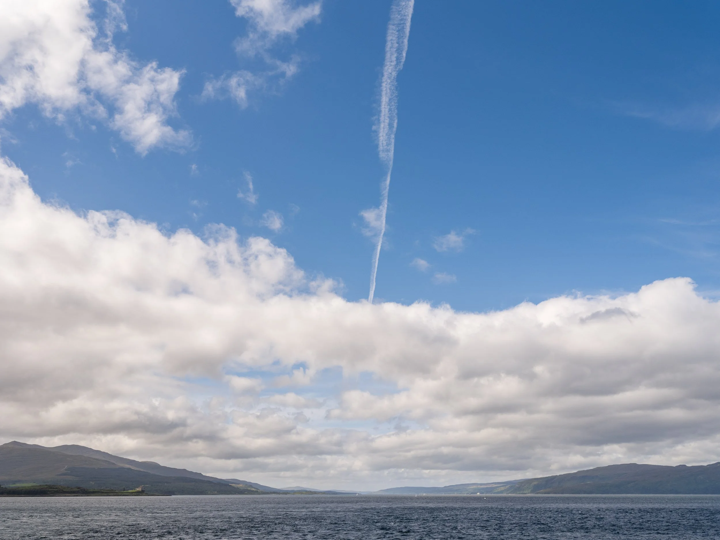 A large body of water with a mountain range in the background under a partly cloudy sky with a contrail from an airplane.