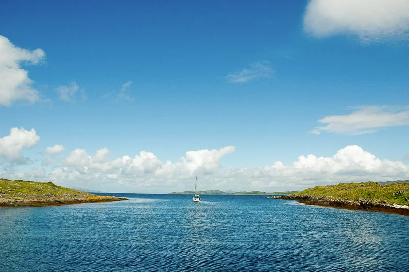 A calm sea with a sailboat in the distance under a partly cloudy sky, flanked by green, rocky shores.