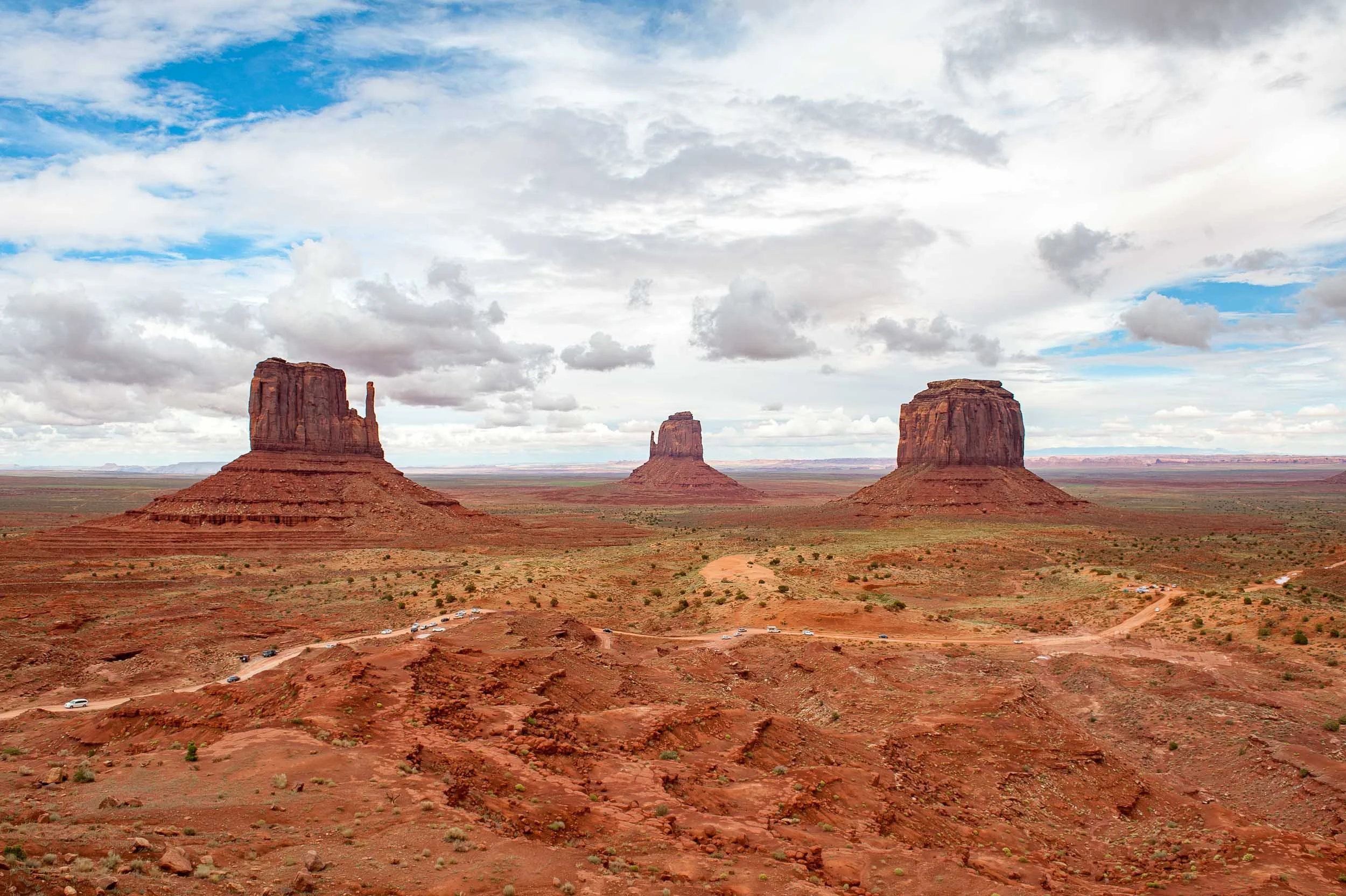 Scenic view of Monument Valley with three large red sandstone buttes under a partly cloudy sky, with a dirt road and parked cars in the foreground.
