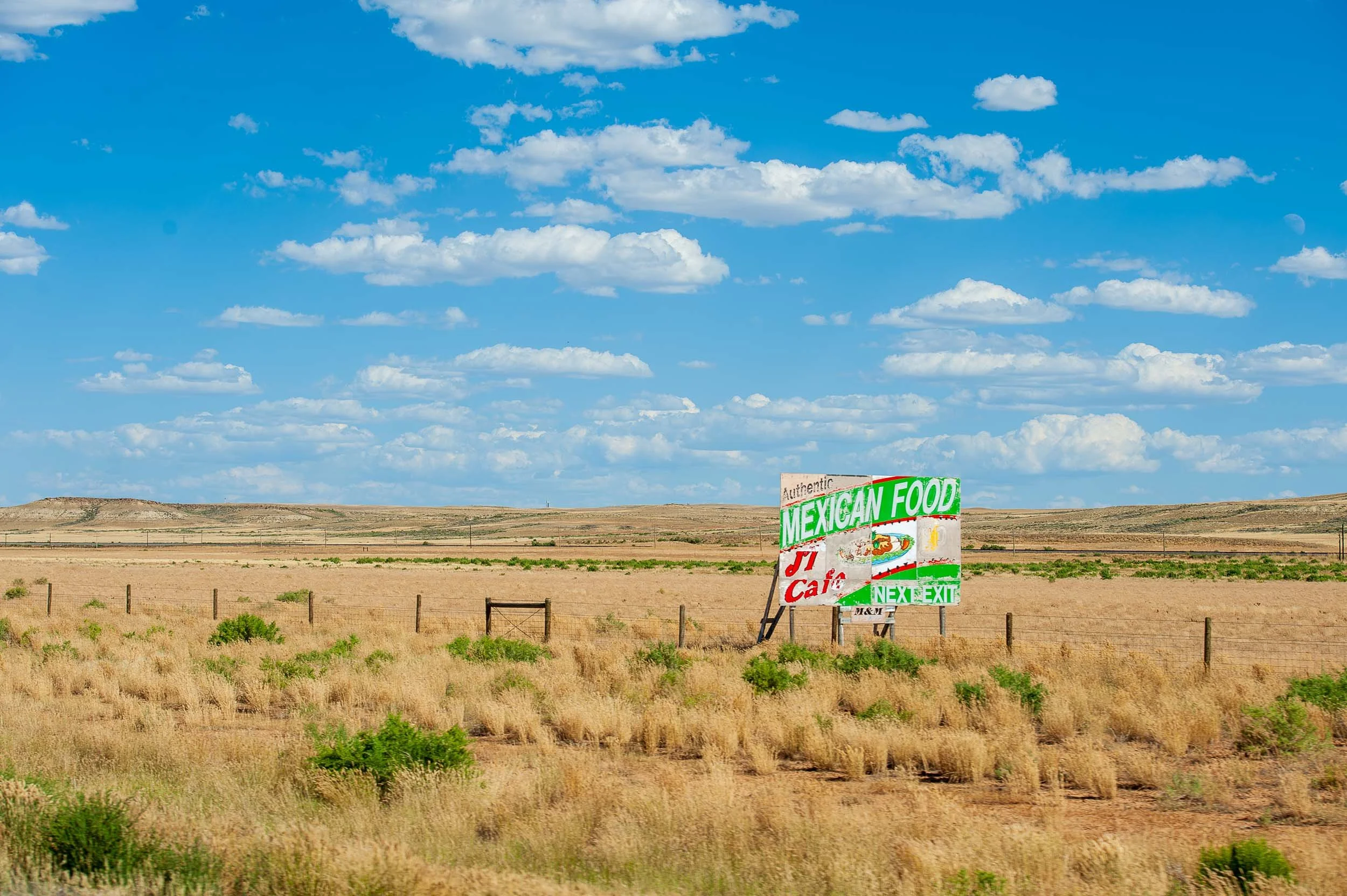 A sign advertising authentic Mexican food and a cafe in a rural, open desert area with dry grass, sparse green bushes, and a chain-link fence under a bright blue sky with scattered clouds.