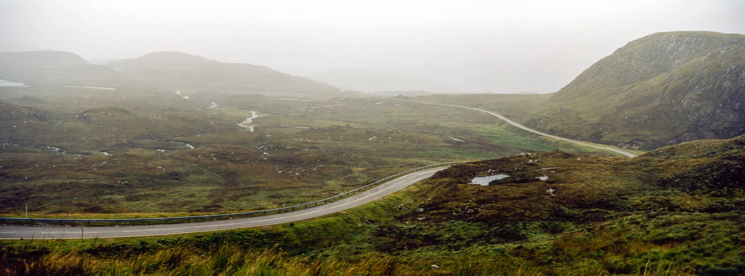 A winding mountain road through a foggy, green and brown landscape with small water pools and rolling hills in the distance.