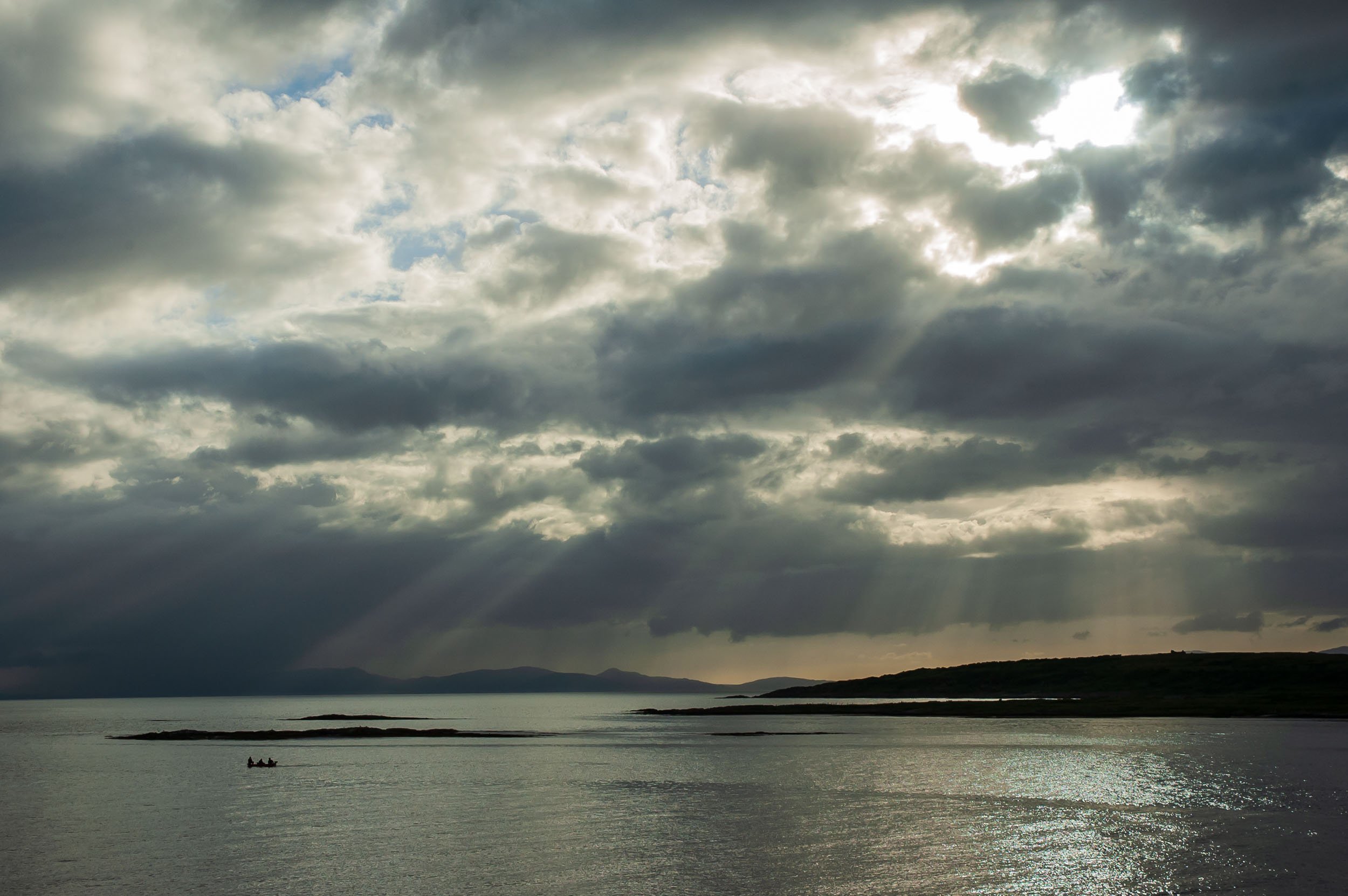 Overcast sky with sun rays breaking through clouds over a body of water with small boat.