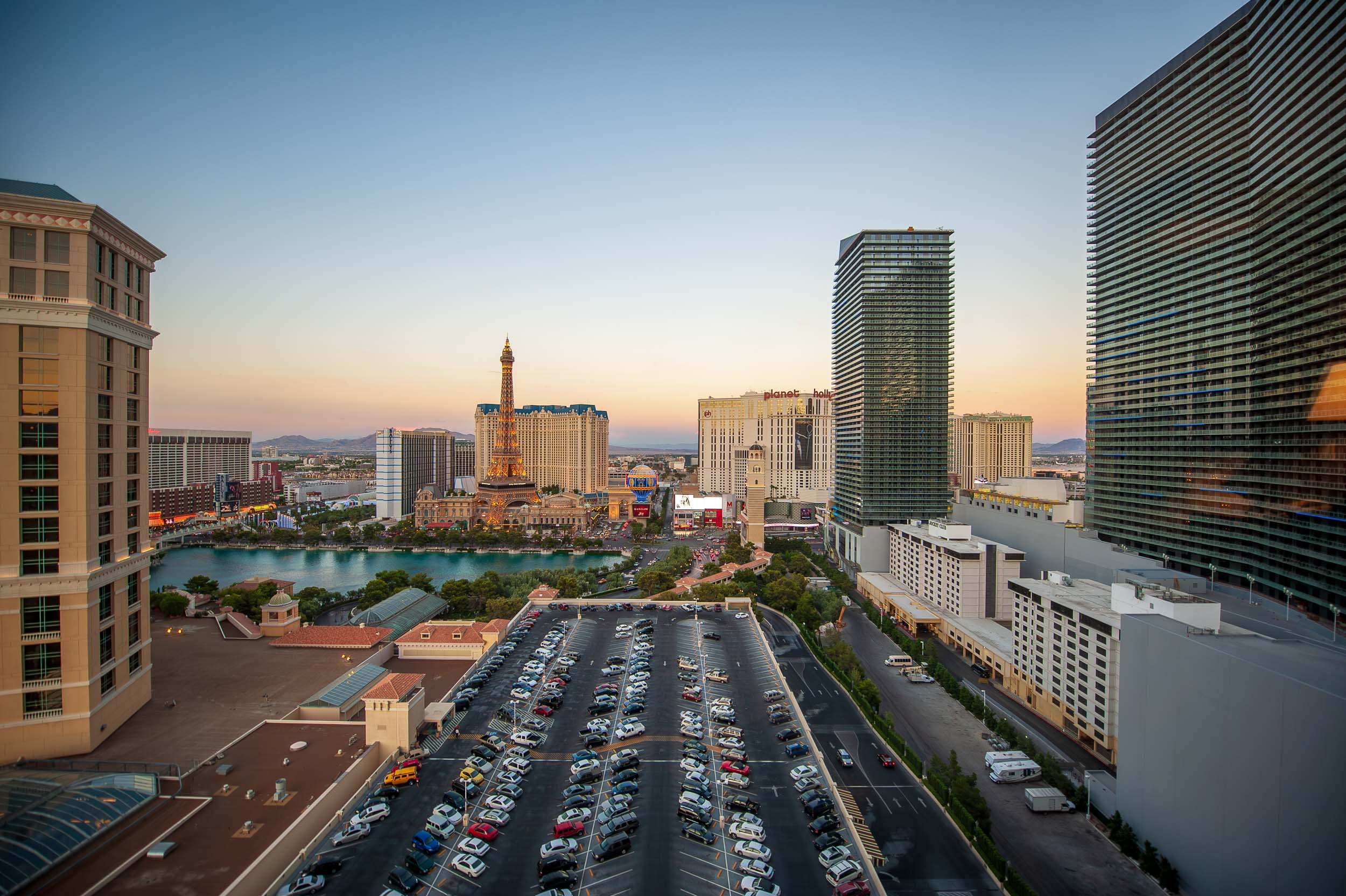 Cityscape of Las Vegas at sunset featuring casino hotels, a lake, and a parking lot filled with cars.