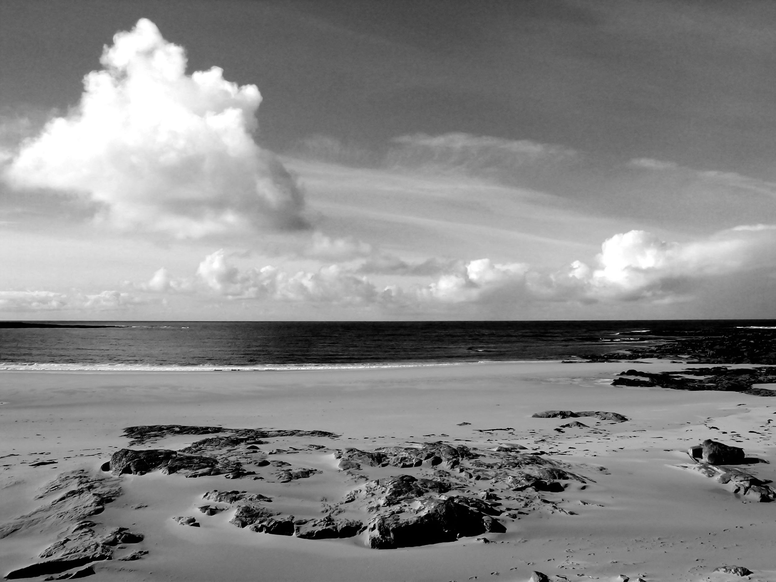 Black and white photo of a beach with rocks in the sand, calm ocean water, and a partly cloudy sky.