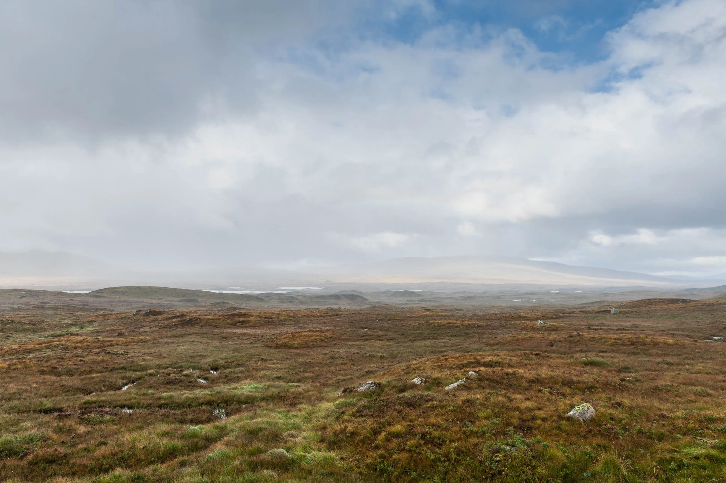 Wide view of a grassy, rocky landscape under a cloudy sky with distant hills.