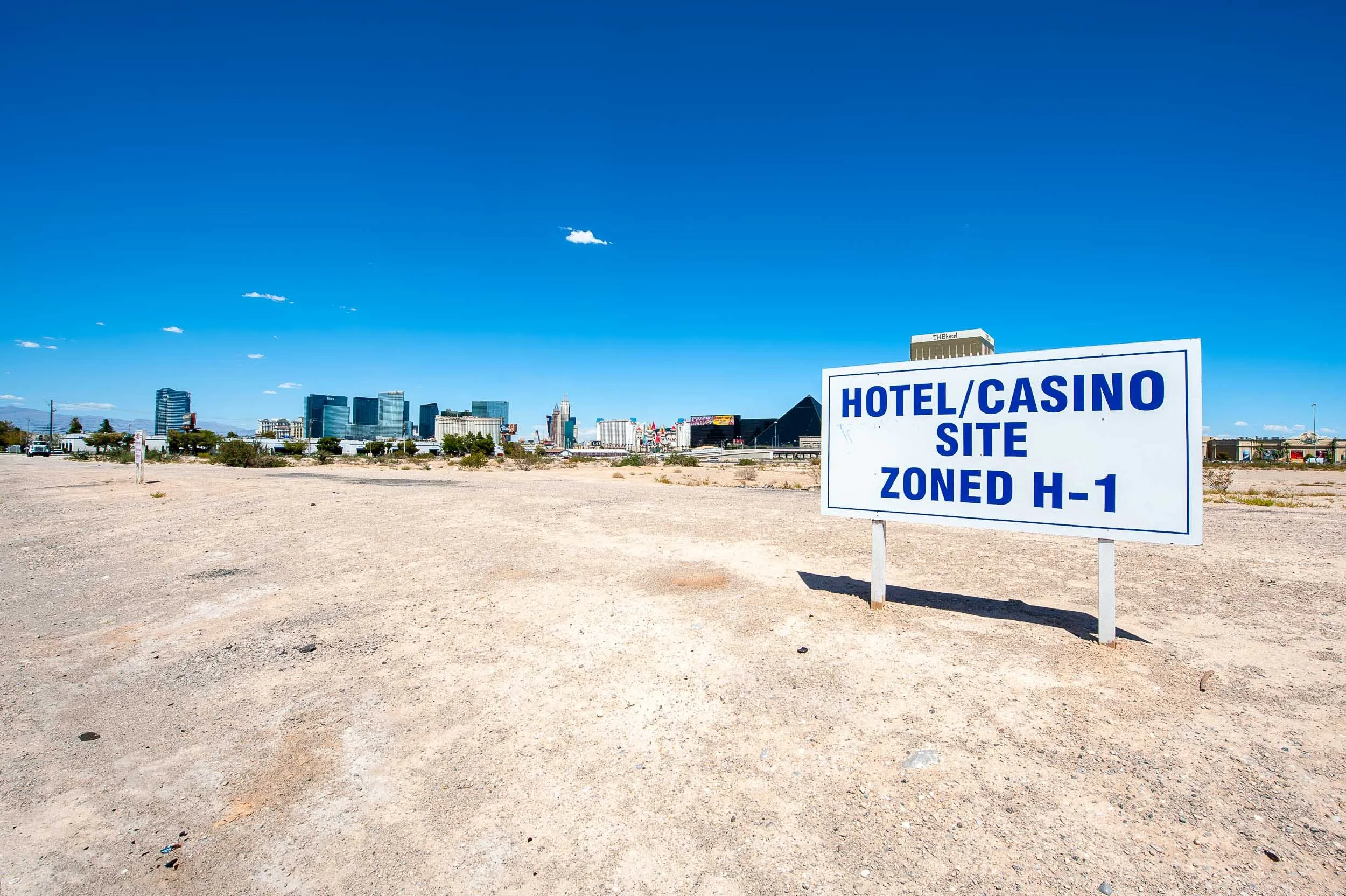 A large white sign with blue text reading "Hotel/Casino Site Zoned H-1" in a desert area with a city skyline in the background under a clear blue sky.