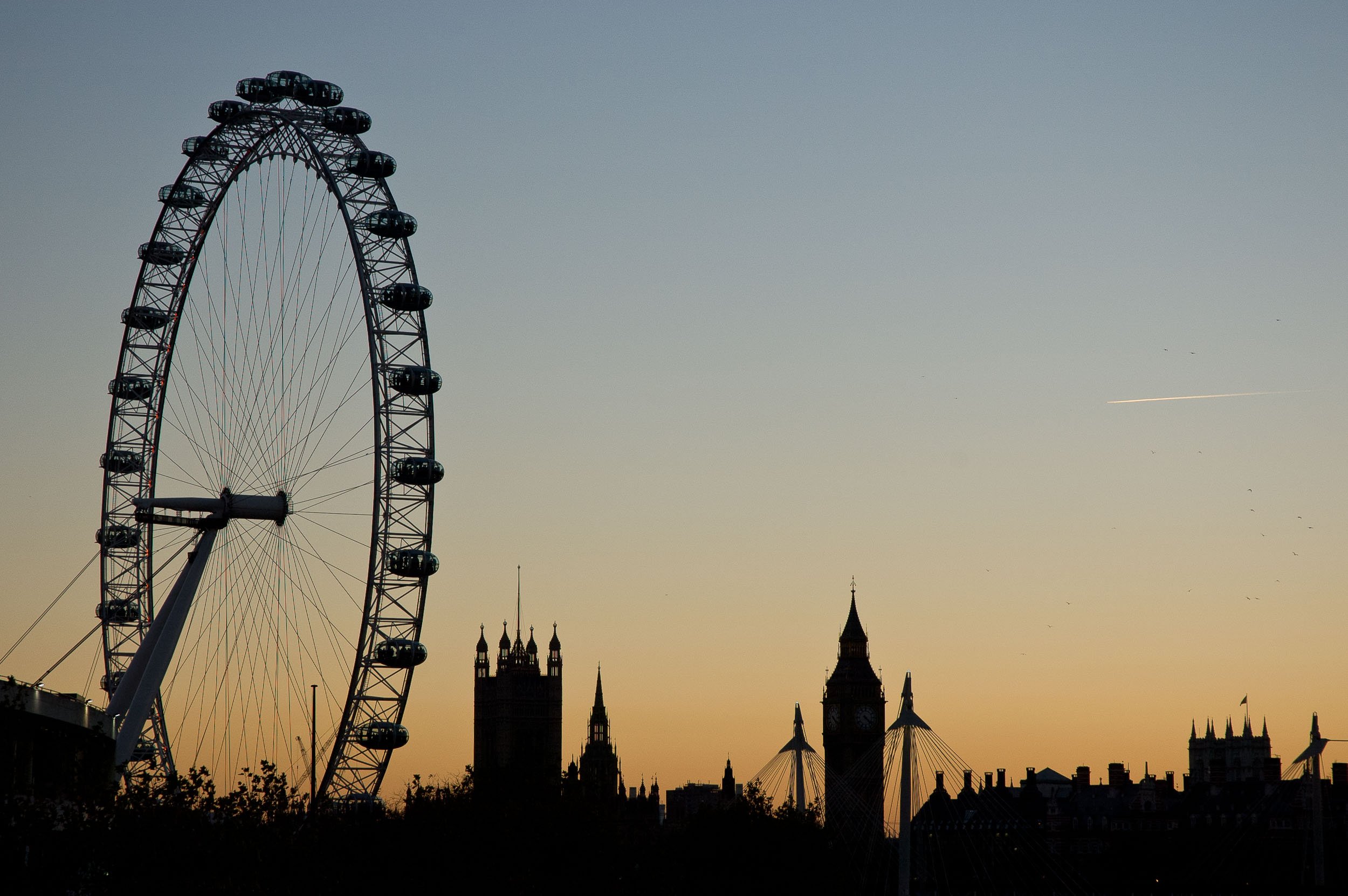 Silhouette of the London Eye Ferris wheel, Big Ben clock tower, and other skyline buildings against a sunset sky.
