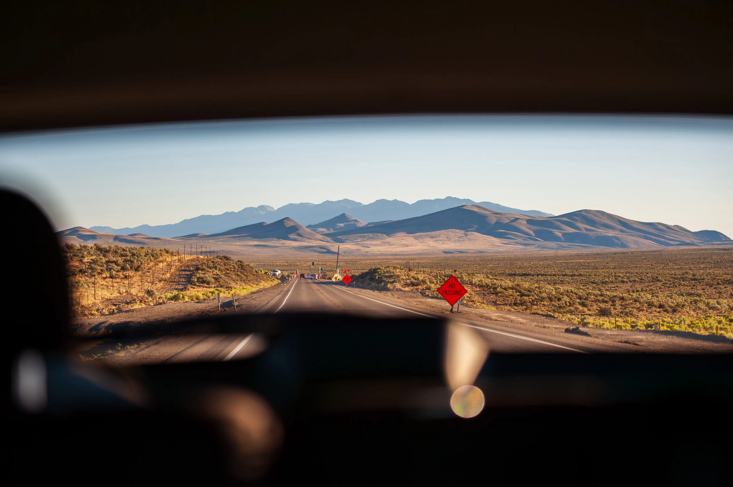 View of a desert highway with mountains in the background, a 'No Passing' road sign on the right, seen from inside a vehicle.