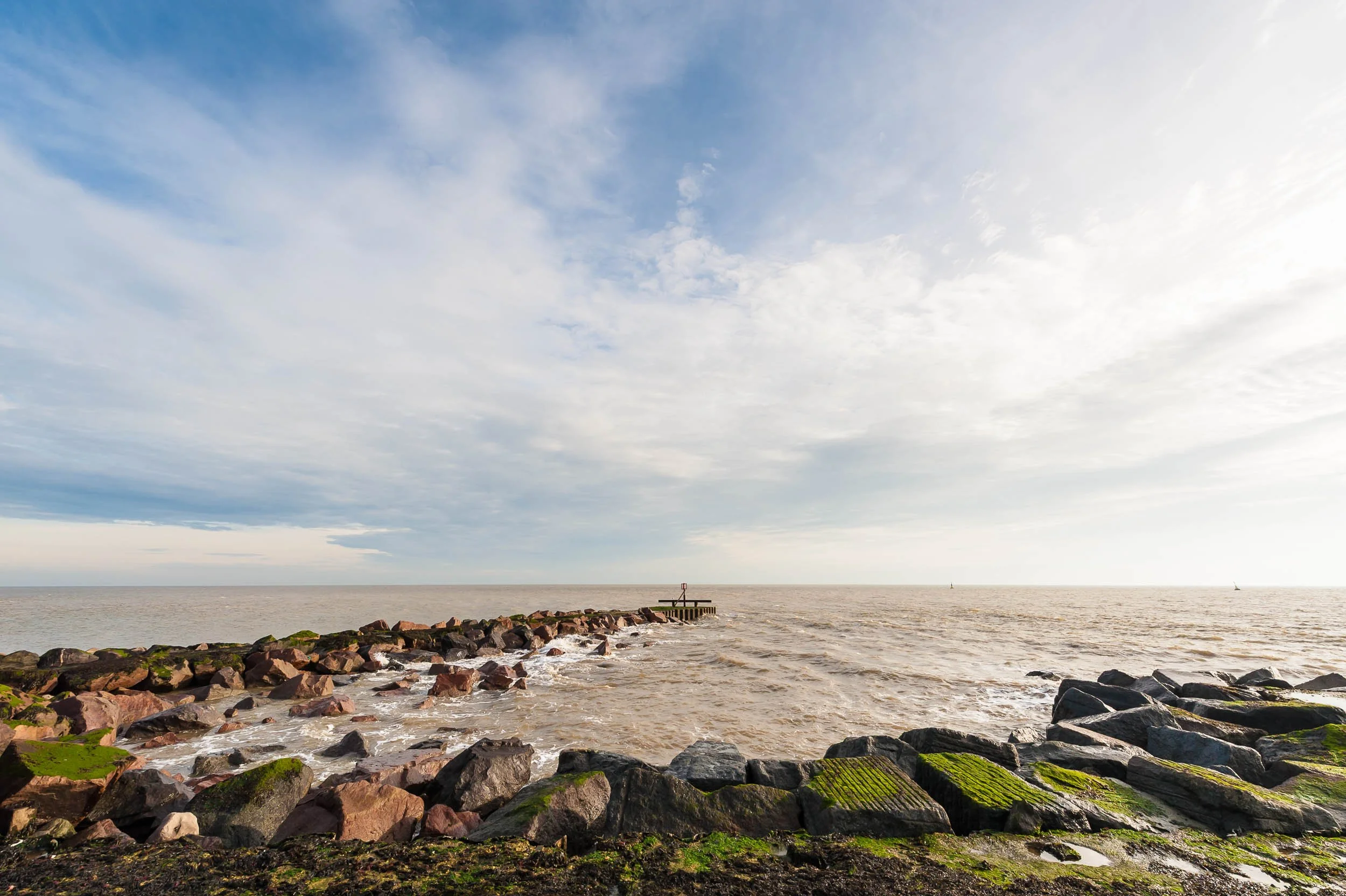 A rocky coastline with moss-covered rocks, leading out to a pier extending into calm ocean water under a partly cloudy sky.