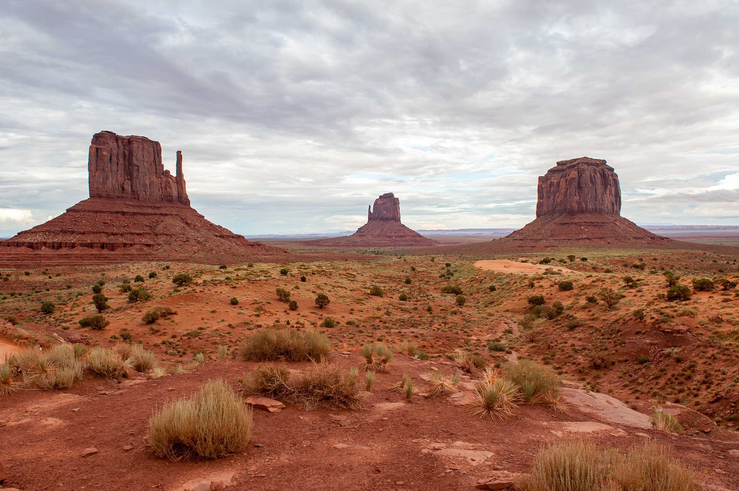 Three large sandstone buttes in Monument Valley desert landscape under overcast sky with sparse desert vegetation.