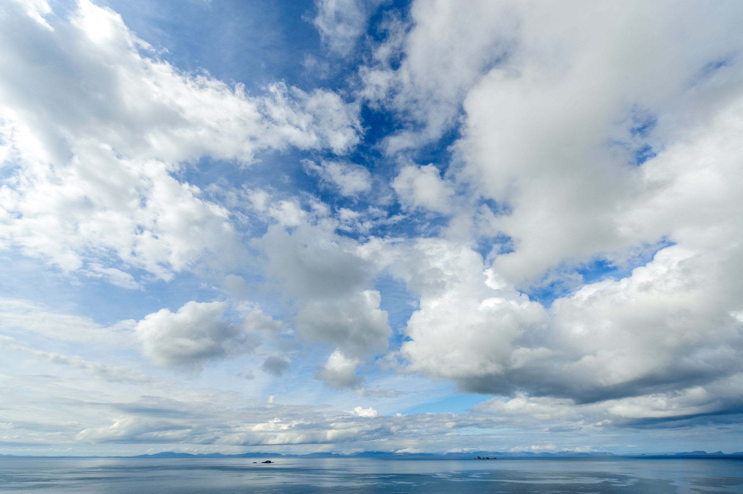 Cloudy sky over calm ocean with distant islands and mountains on the horizon.