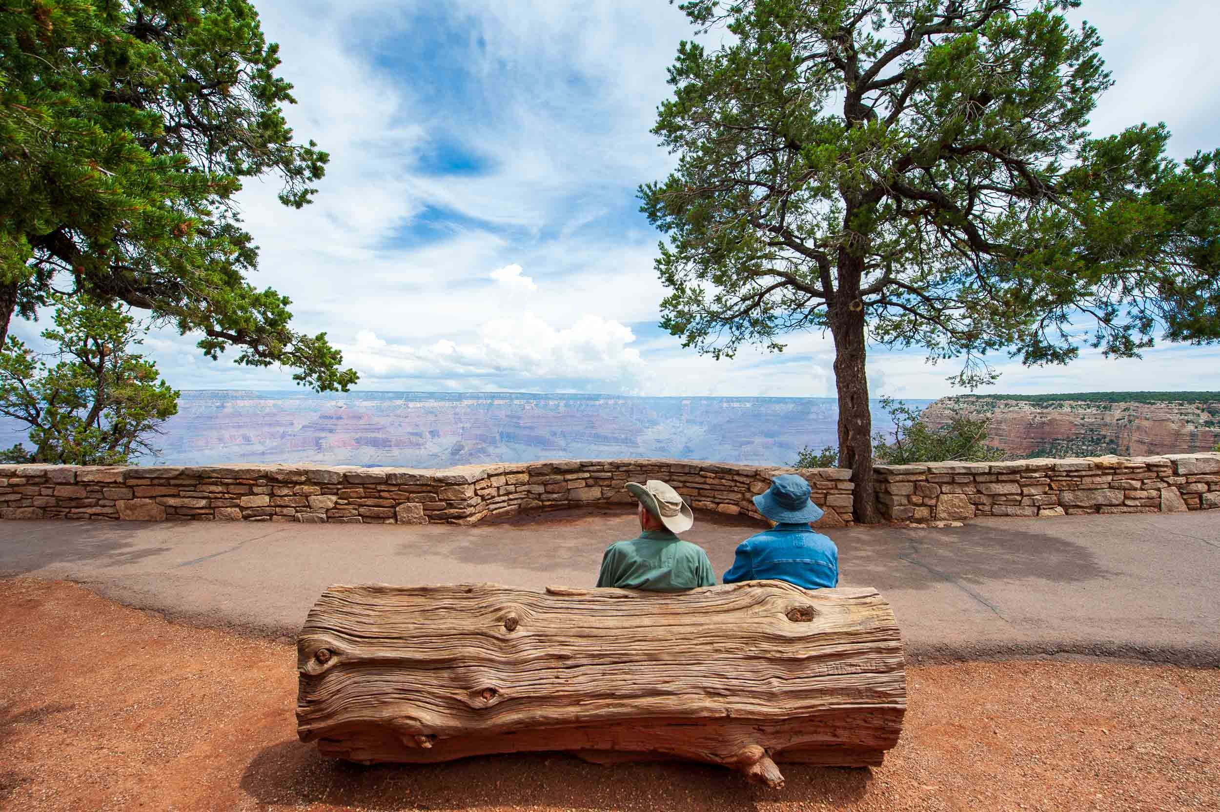 Two people sitting on a wooden bench overlooking the canyon at Grand Canyon National Park, with trees and a stone wall in the background.