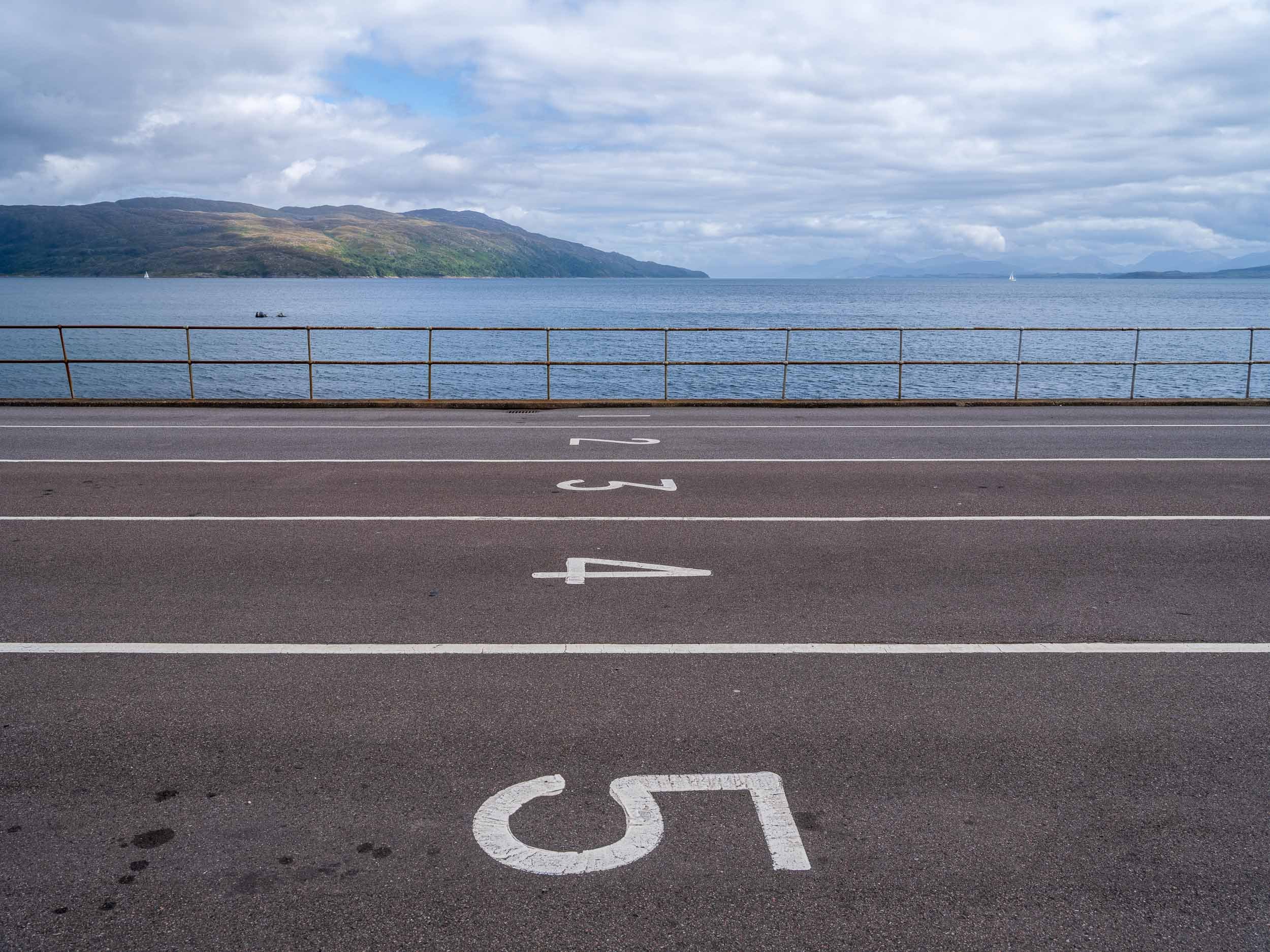 Empty numbered parking spaces 4 and 5 in front of a body of water with mountains in the background, and a cloudy sky.