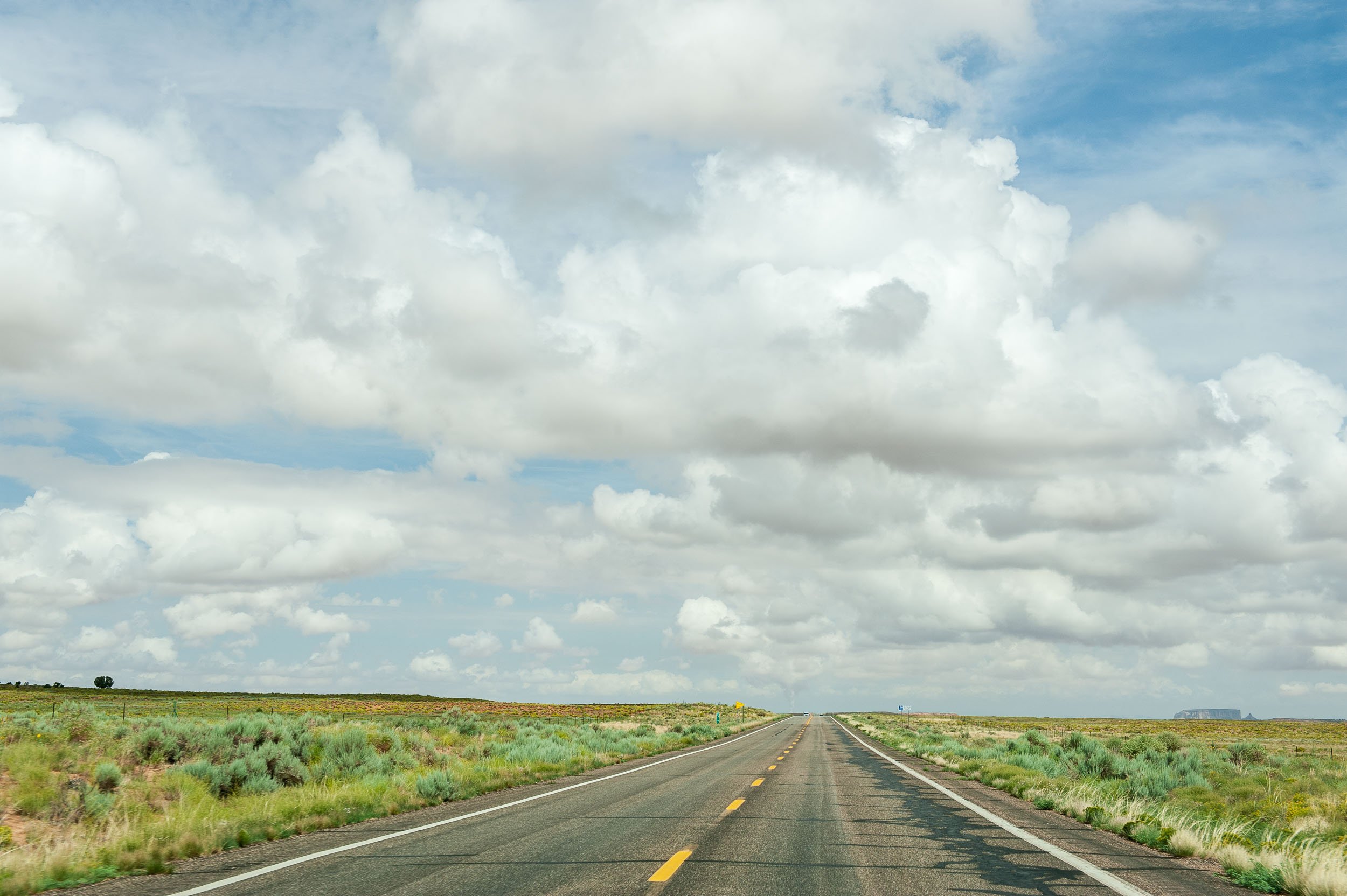 A long empty road stretches into the horizon through a flat, open landscape under a partly cloudy sky.
