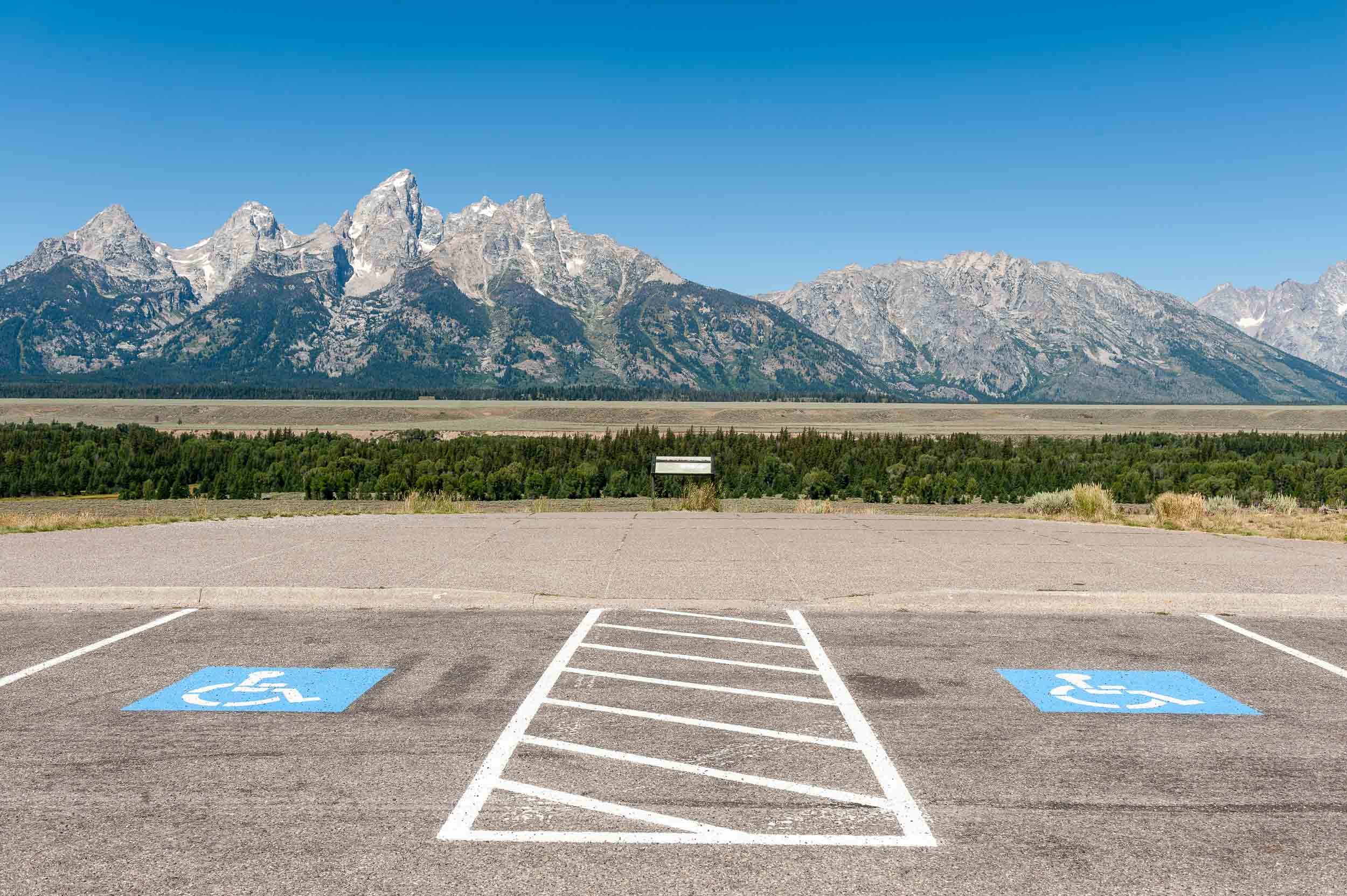 Empty parking lot with two accessible parking spaces in the foreground, mountains and forest in the background, clear blue sky.