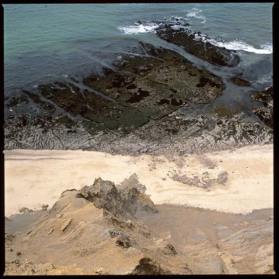 A rocky coastline with dark rocks in the ocean and light sandy beach in the foreground.