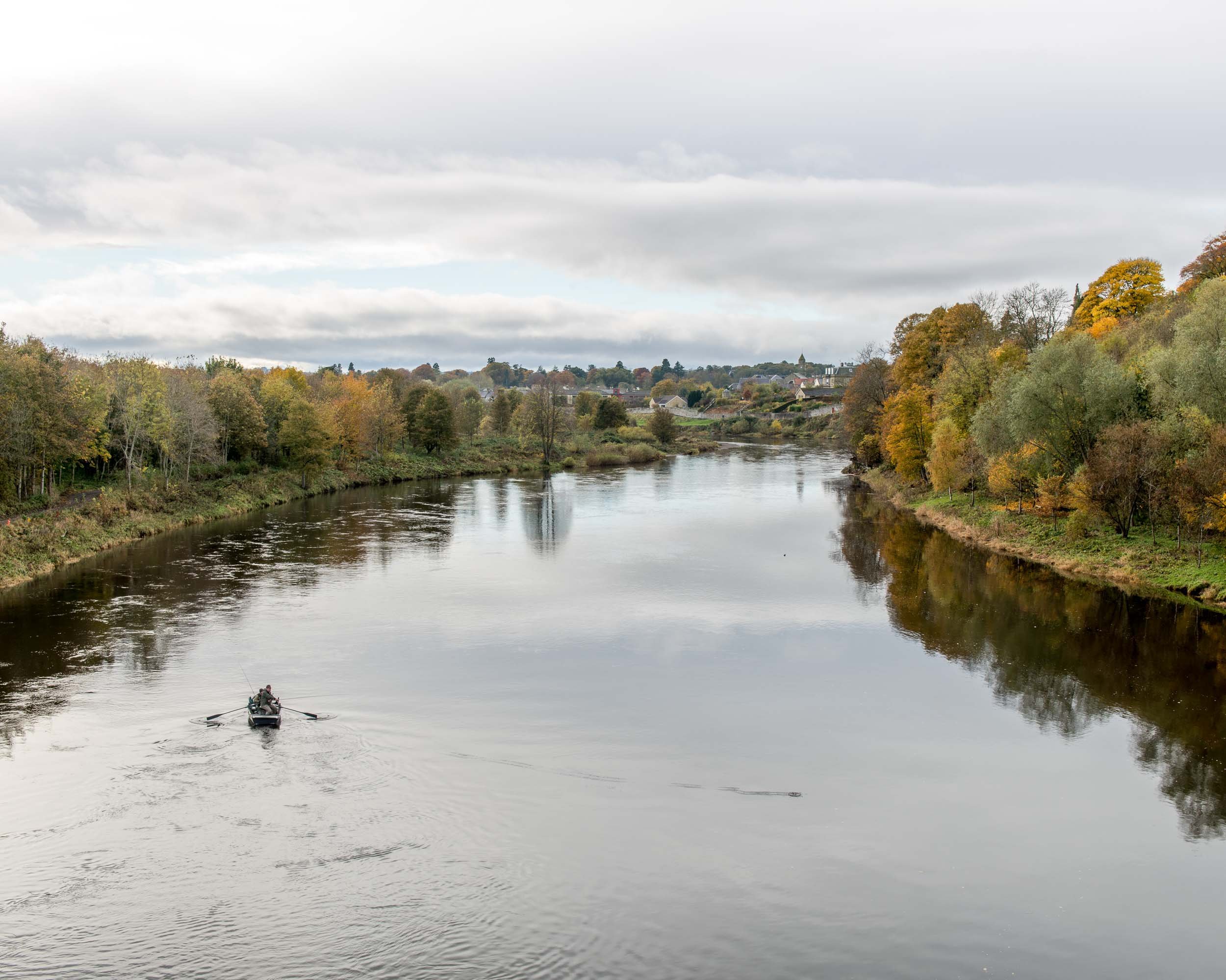 A calm river flowing through a landscape with trees on either side, reflecting the cloudy sky above. A small boat with two people is seen in the river.