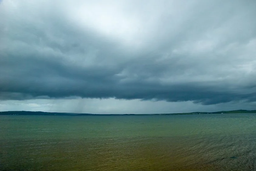 Dark storm clouds over a body of water, possibly a lake or sea, with land visible in the distance during overcast weather.