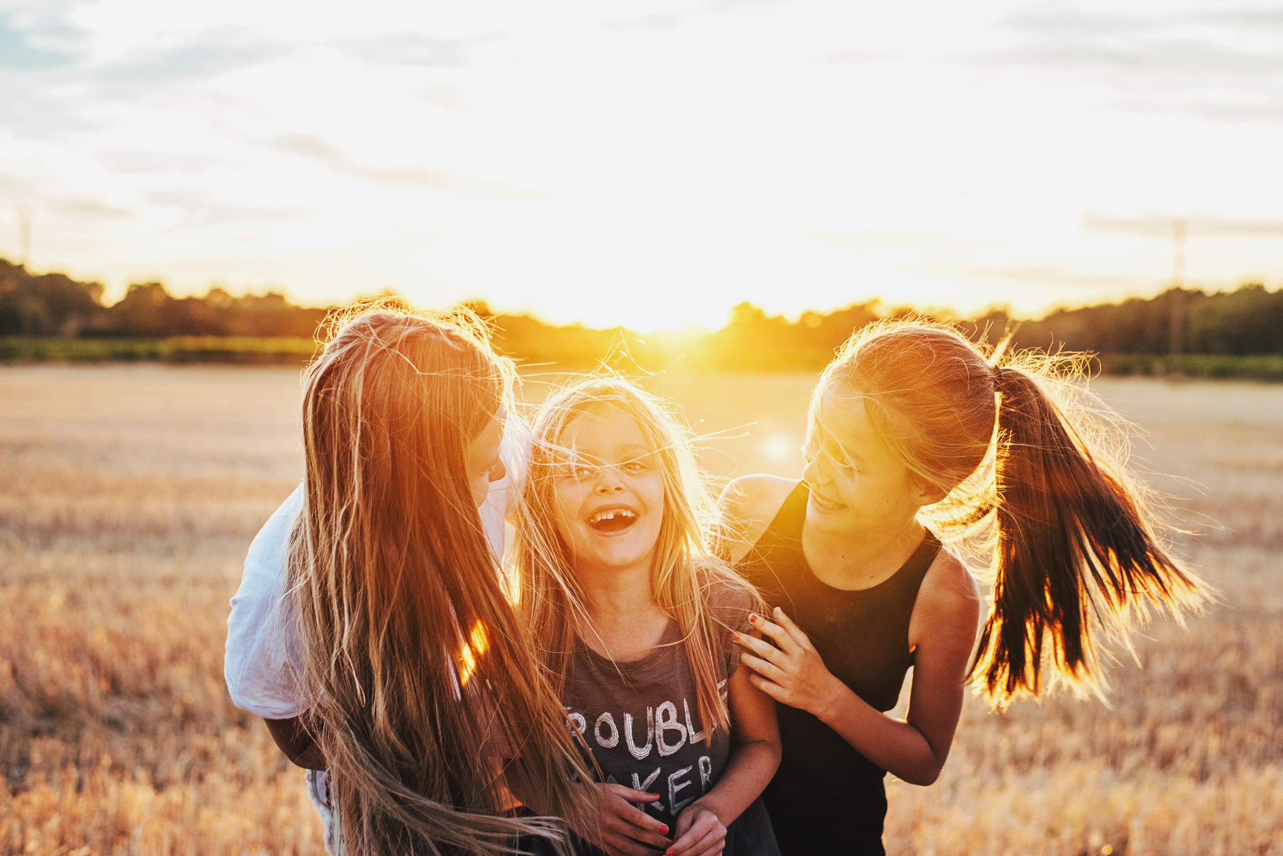 Three girls stand in cornfield laughing at sunset Essex UK Documentary Portrait Photographer