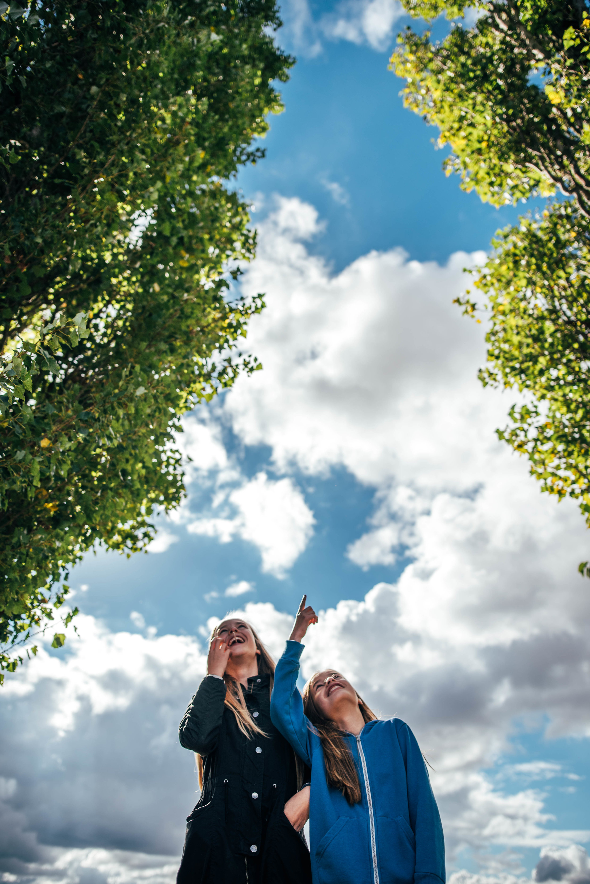 Two girls stand framed by trees against big cloudy sky Essex UK Documentary Portrait Photographer