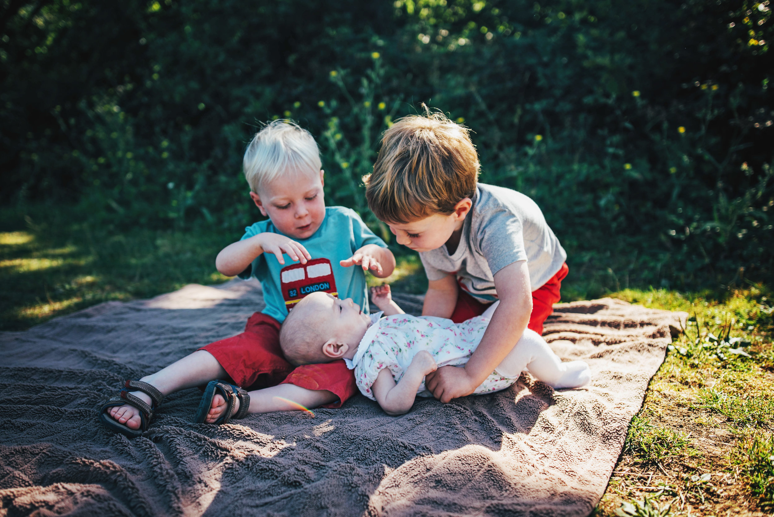 Two big brothers hug baby sister in sunny park Essex UK Natural Documentary Portrait Photographer