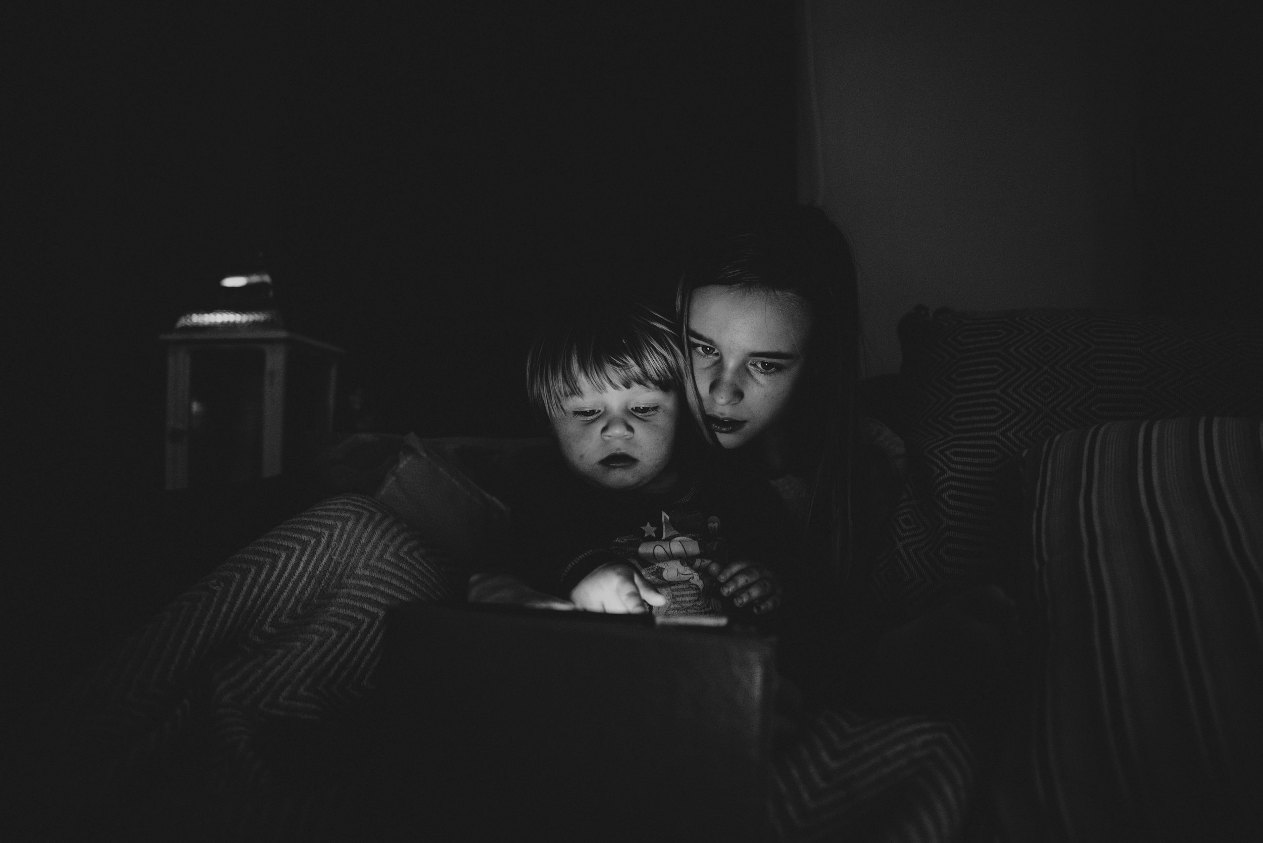 Girl and baby boy look at iPad in the dark Essex UK Documentary Photographer
